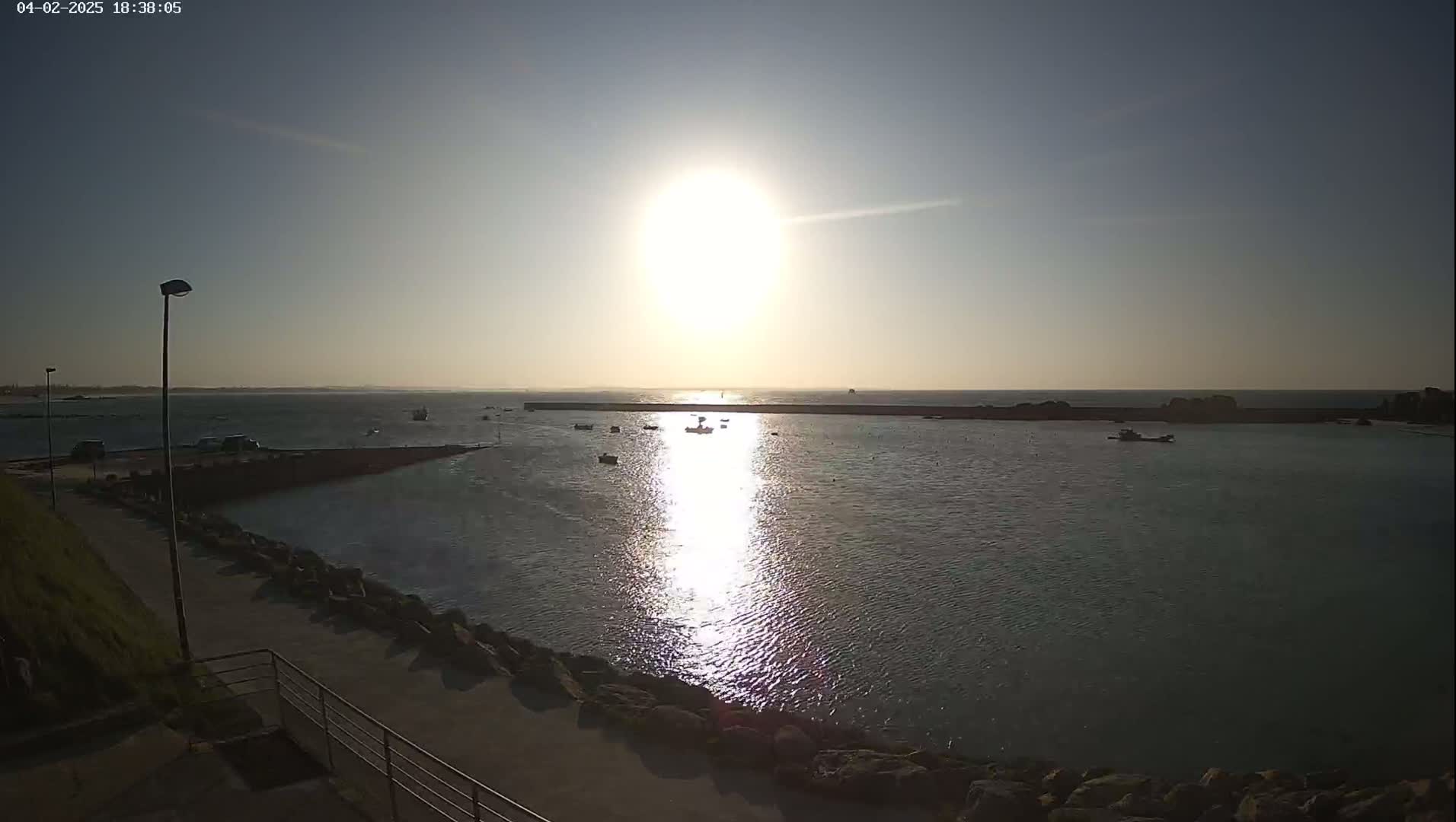 A bright sun shines over a calm harbor with several small boats, a paved walkway, and rocky shoreline under a clear sky.