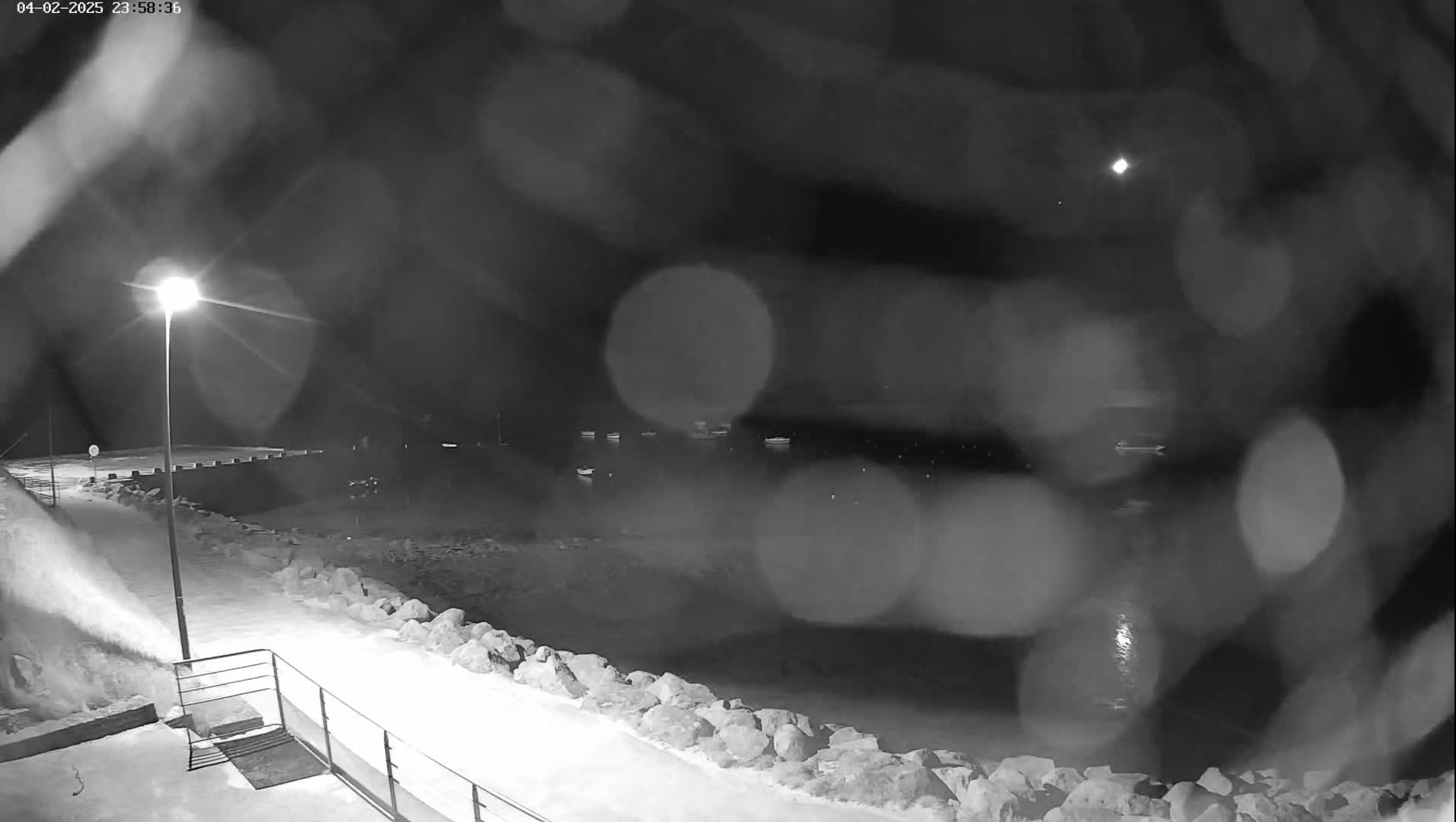 A nighttime, snowy scene shows a snow-covered walkway along a rocky shoreline with several boats visible in the dark water beyond, under a rain- or snow-blurred sky.