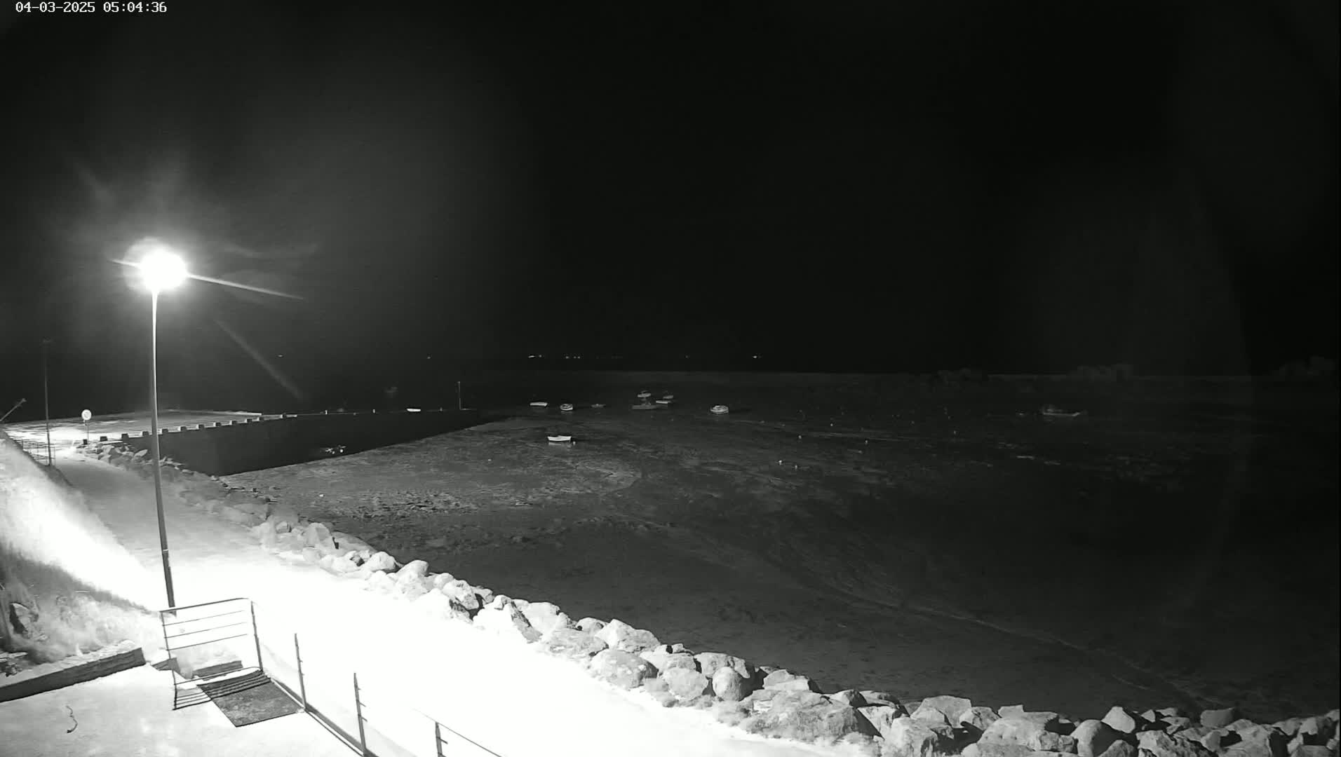 A snow-covered harbor at night, illuminated by a single lamppost, shows several small boats at rest in the water.