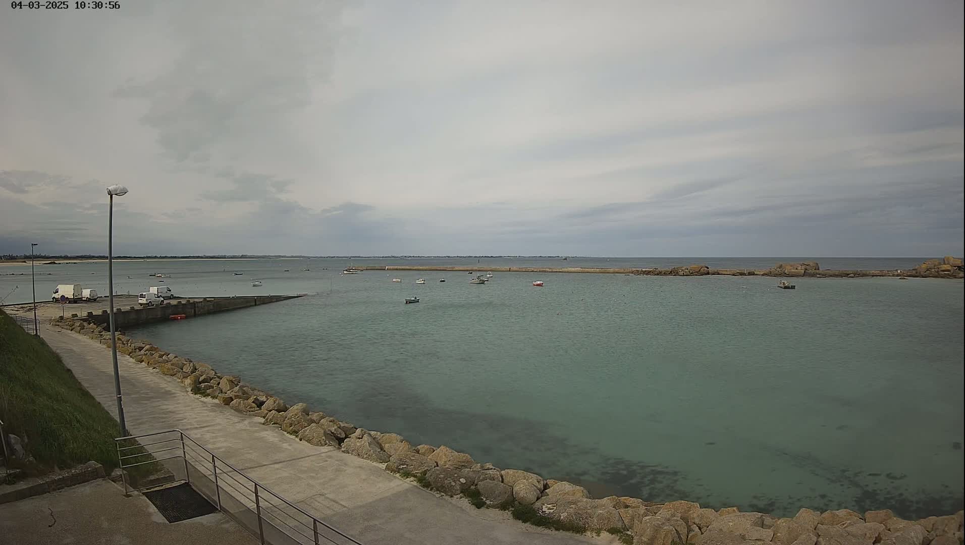 A calm, overcast day reveals a small harbor with several boats, a paved walkway lined with rocks, and a few vehicles parked nearby.