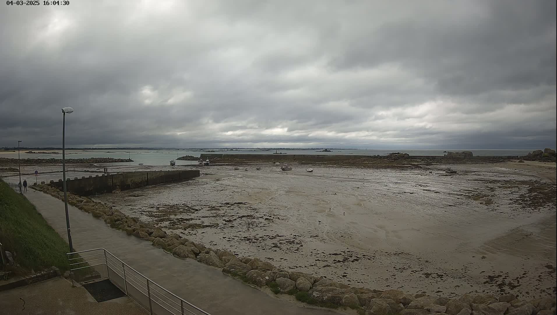 A mostly cloudy day reveals a low tide exposing a large expanse of mudflats in a harbor with several small boats, a walkway, and a distant coastline.