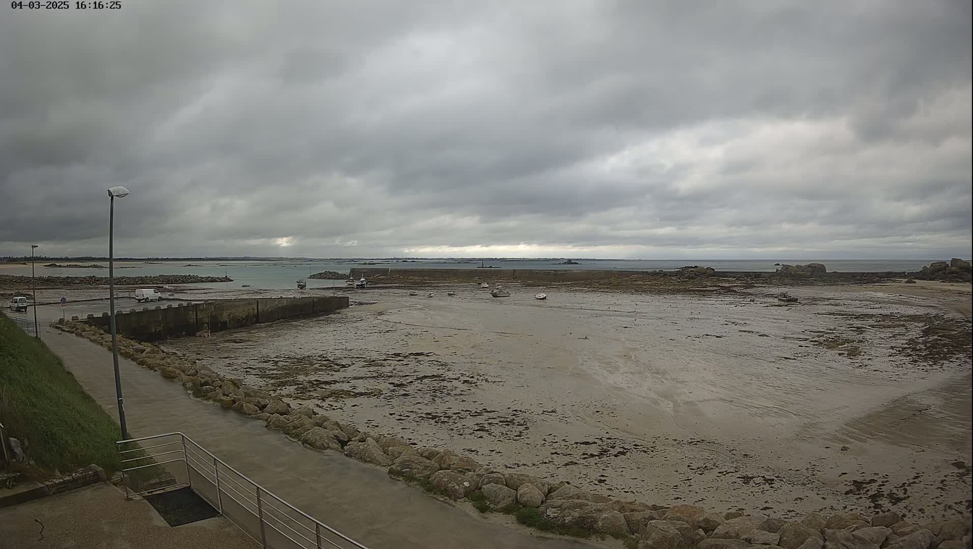 A mostly overcast sky hangs over a harbor, where a mostly empty, muddy tidal flat is exposed with several small boats and a few vehicles visible near a low stone wall.