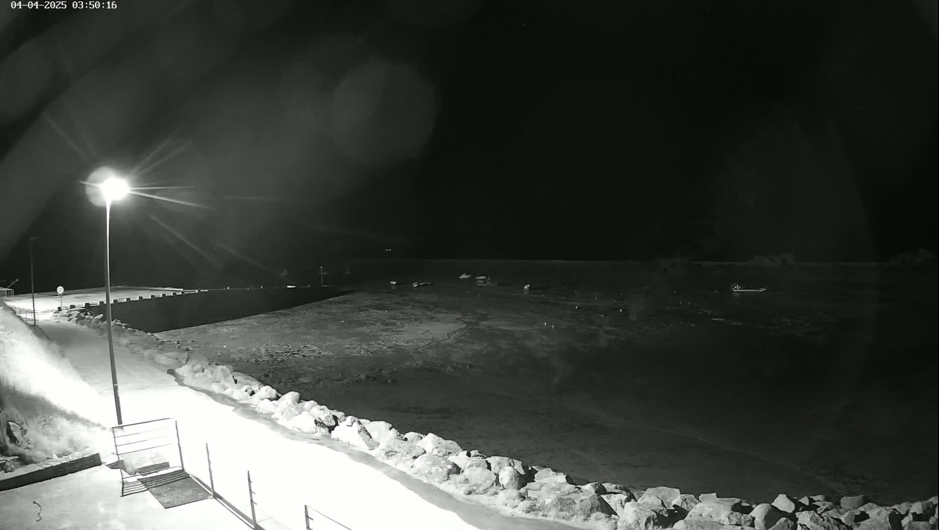 A nighttime, black and white view of a harbor with several boats, a snow-covered walkway, and a lit lamppost.
