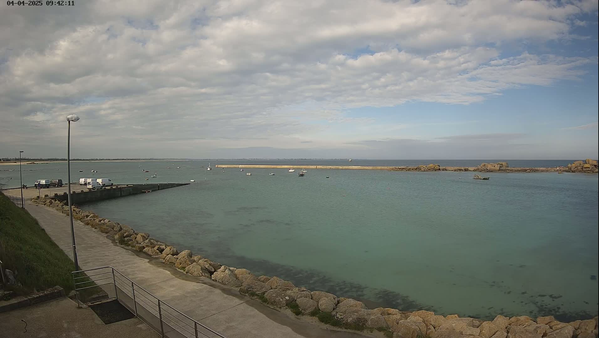 A calm, partly cloudy day reveals a harbor with several small boats, a concrete walkway lined with rocks, and a distant sandy shore.