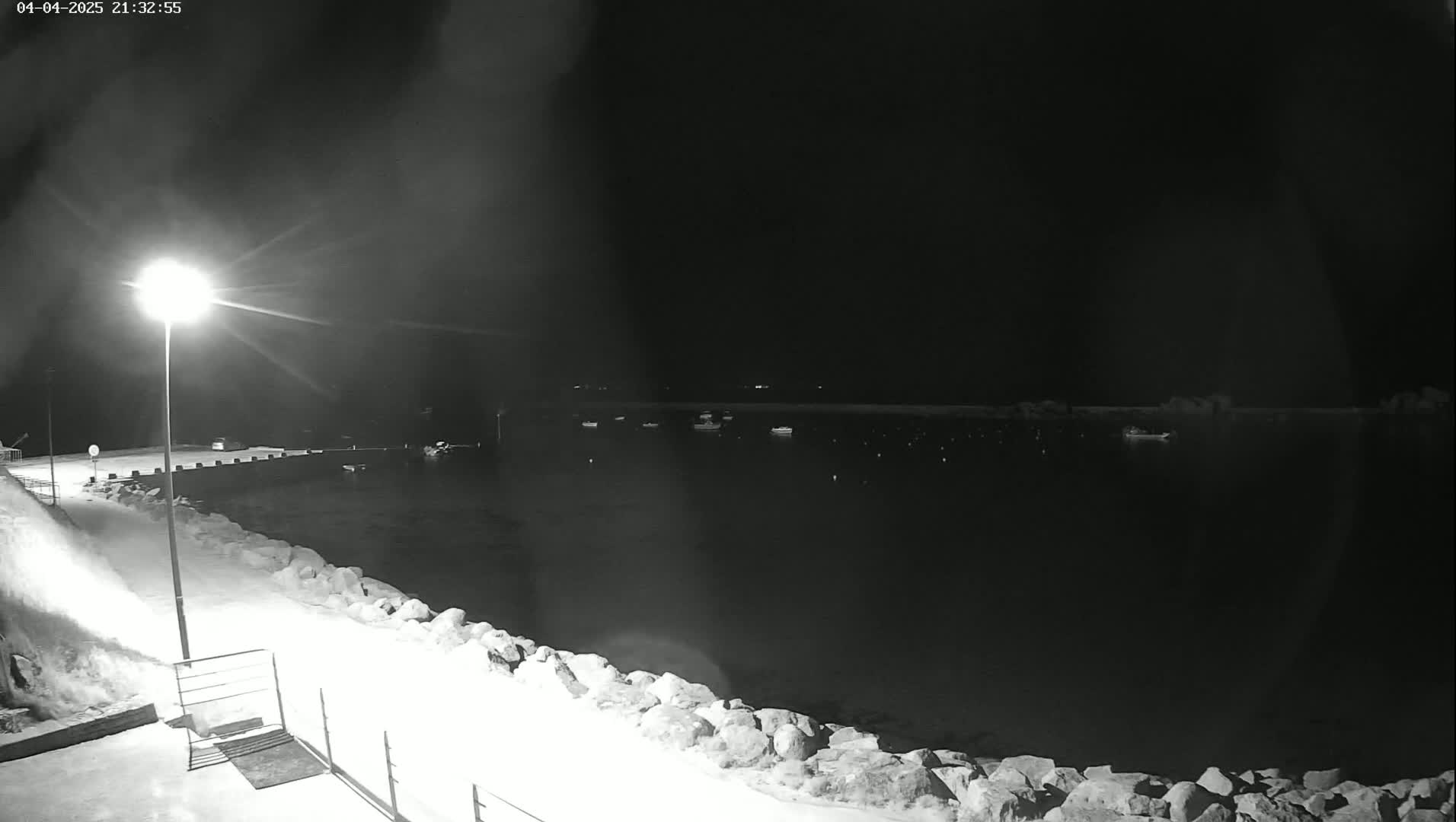 A nighttime, snow-covered harbor scene shows several boats moored in dark water under a light pole.