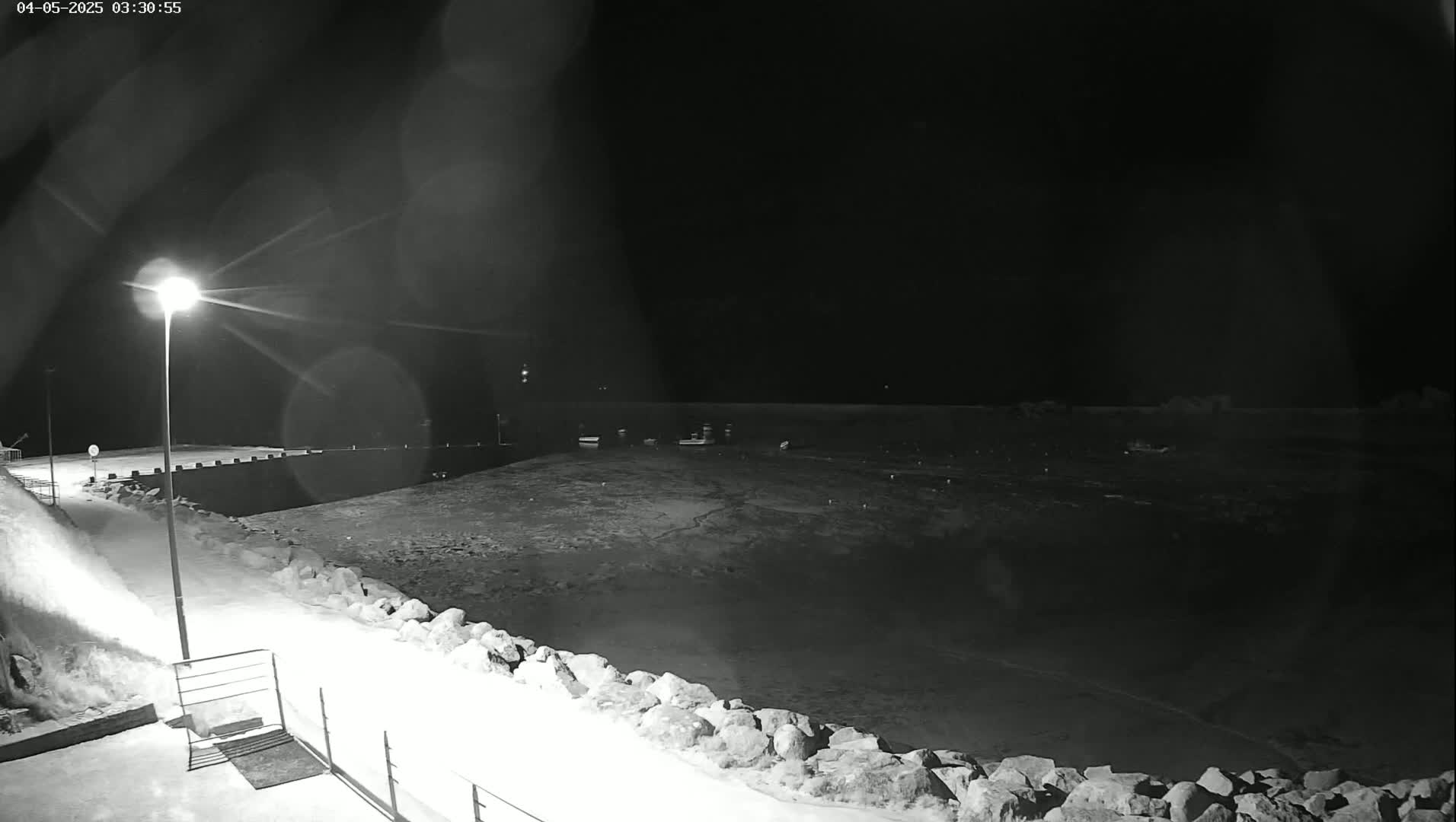A nighttime, snow-covered harbor scene shows a few boats at rest in the dark water, illuminated by a single lamppost on the shoreline.