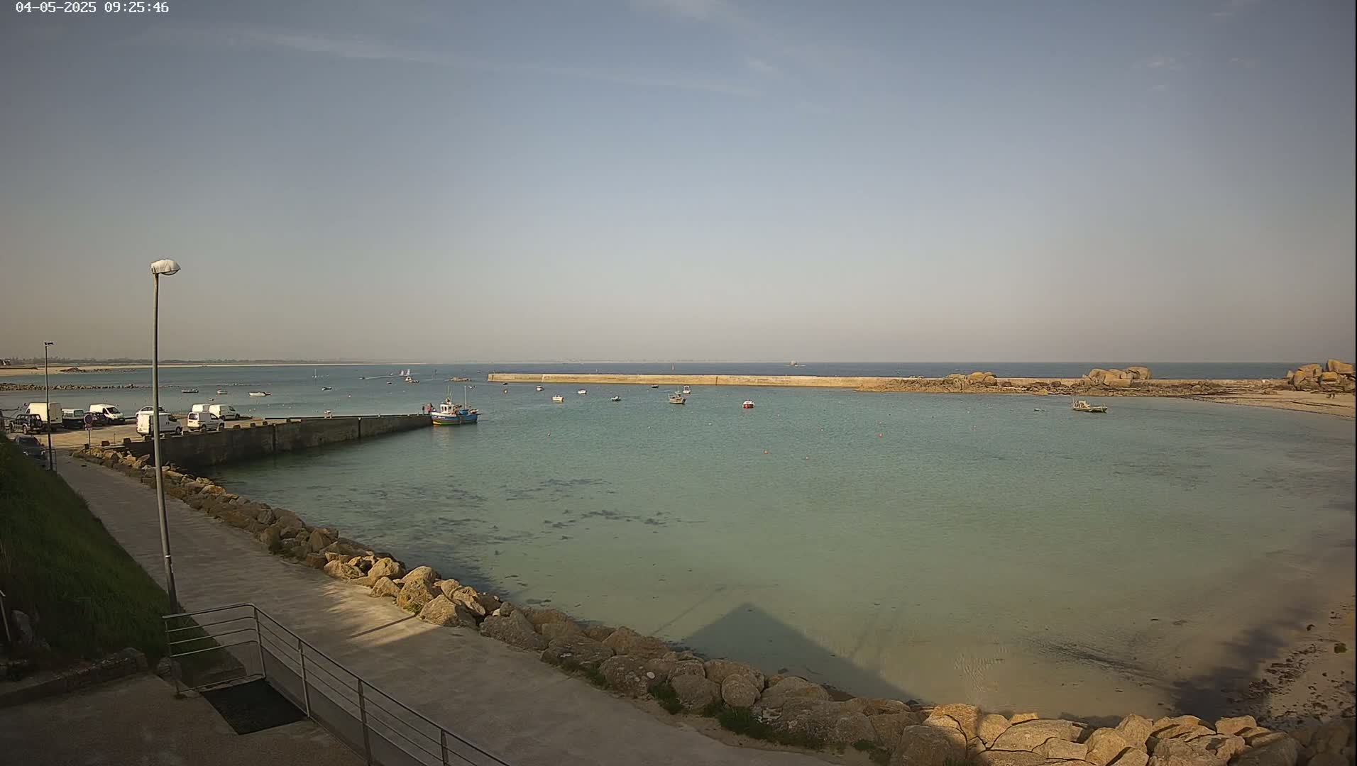 A calm, sunny day shows a harbor with several small boats, a long stone breakwater, and a paved walkway lined with rocks.