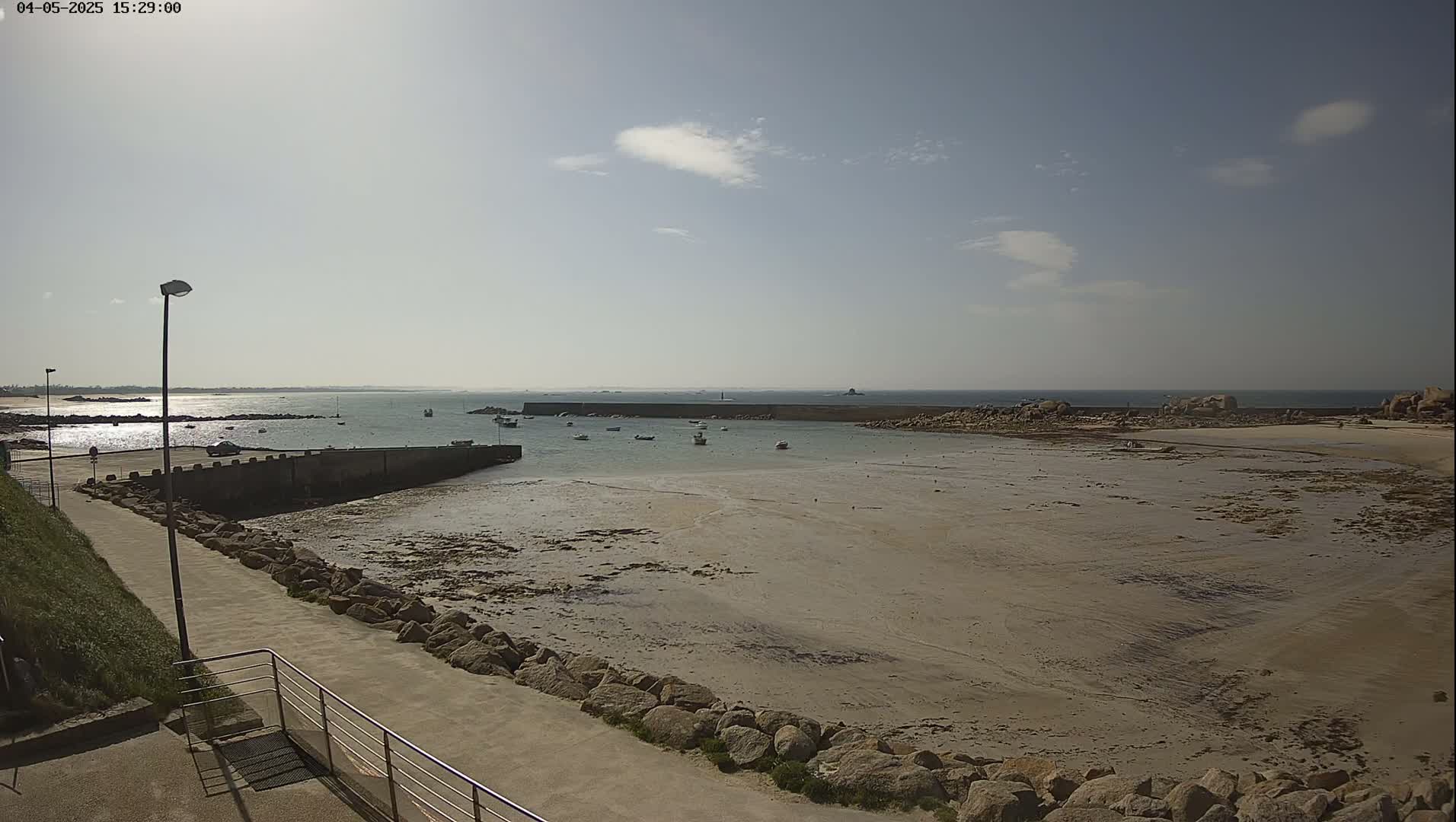 A mostly sunny day shows a rocky shoreline with a small harbor containing several boats at low tide, a paved walkway leading to the water, and a distant view of the ocean.