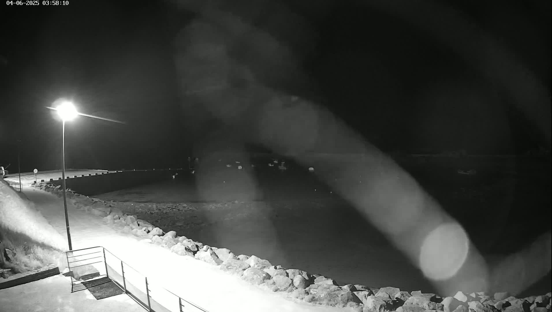 A nighttime, snow-covered waterfront scene shows a body of water with several indistinct objects, a stone seawall, and a lit streetlamp.