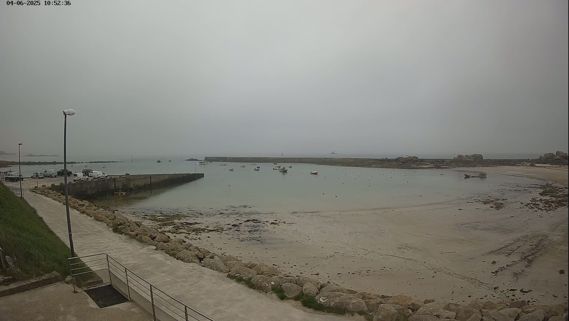 A small harbor with several boats is visible on a foggy day, next to a sandy beach with rocks and a paved walkway leading away from the scene.