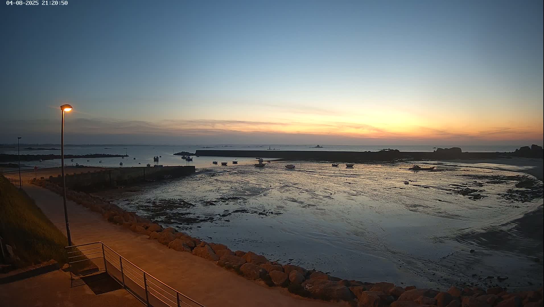 A tranquil harbor scene at sunset shows several small boats resting in shallow, mud-flat waters, partially enclosed by a low stone wall, under a clear, twilight sky.