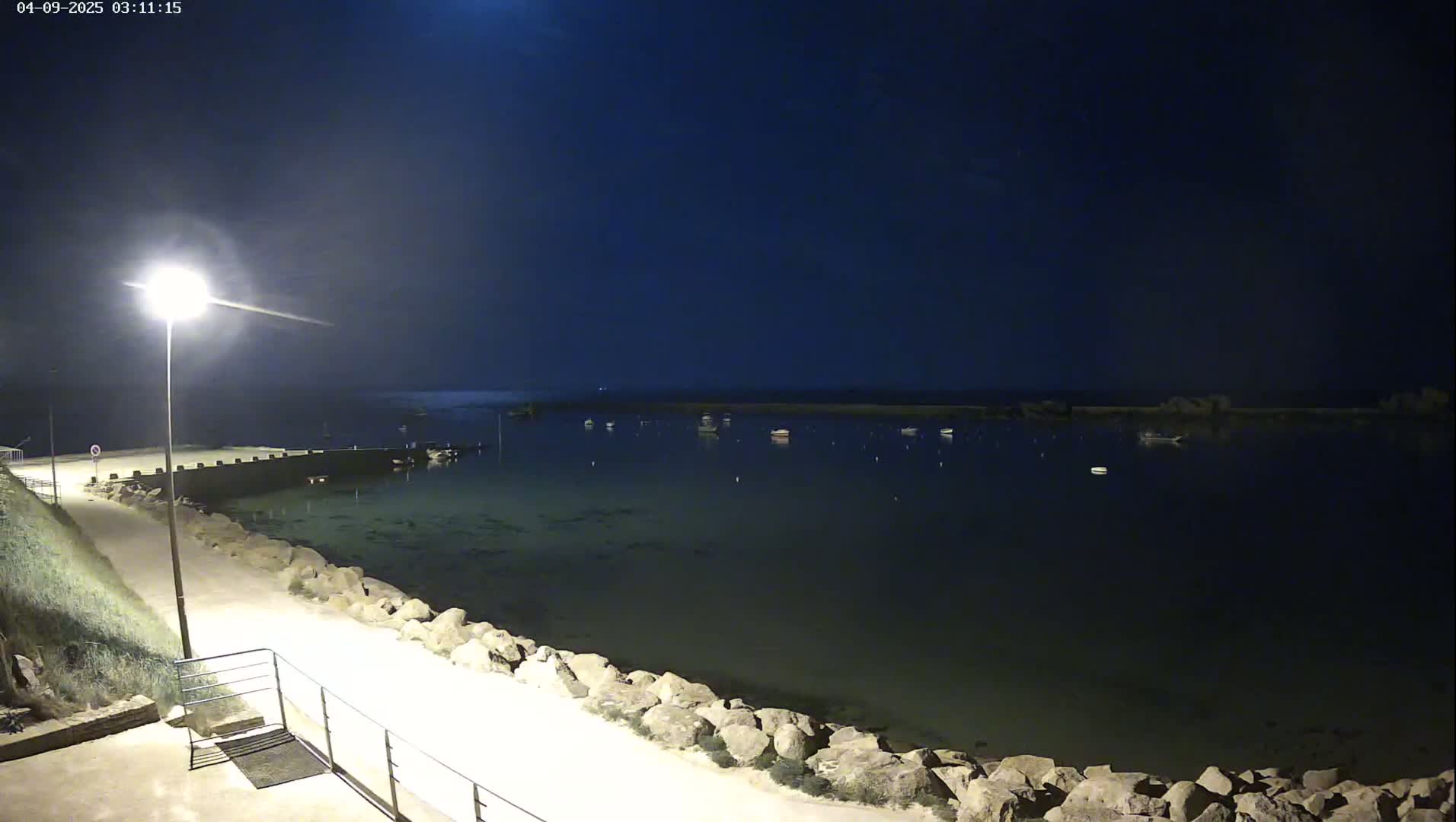 A nighttime view of a calm body of water with several boats moored, a lit walkway along a rocky shoreline, and a single streetlight illuminating the scene.
