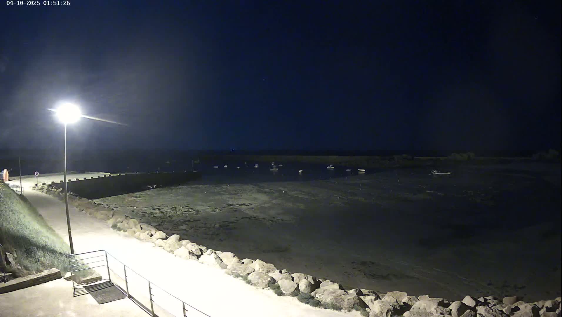 A nighttime view of a calm harbor with several small boats at low tide, illuminated by a single lamppost on a paved walkway beside a rocky shoreline.