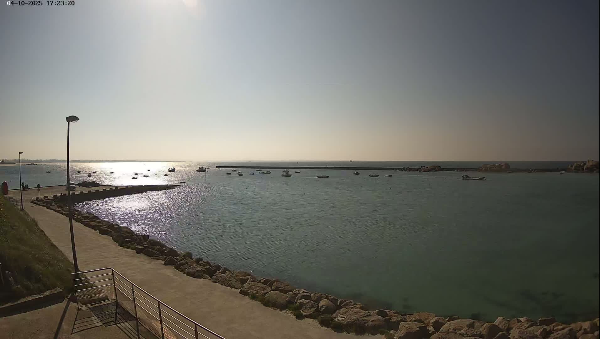 A sunny day reveals a calm harbor with several small boats, a paved walkway, and a rocky shoreline.