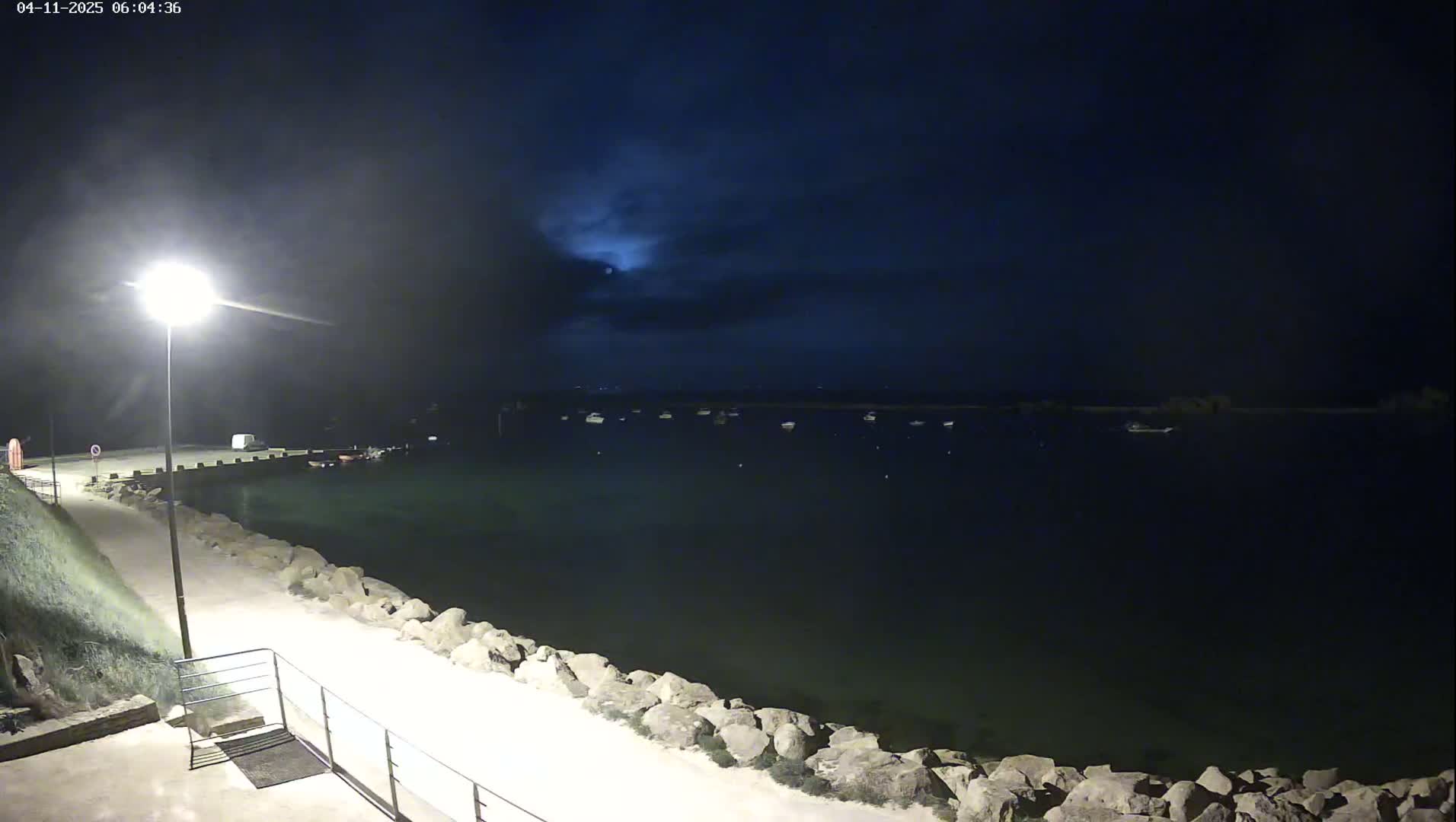 A moonlit nighttime scene shows a calm body of water with several boats at anchor, a paved walkway lined with rocks leading to a pier, and a single lamppost illuminating the path.