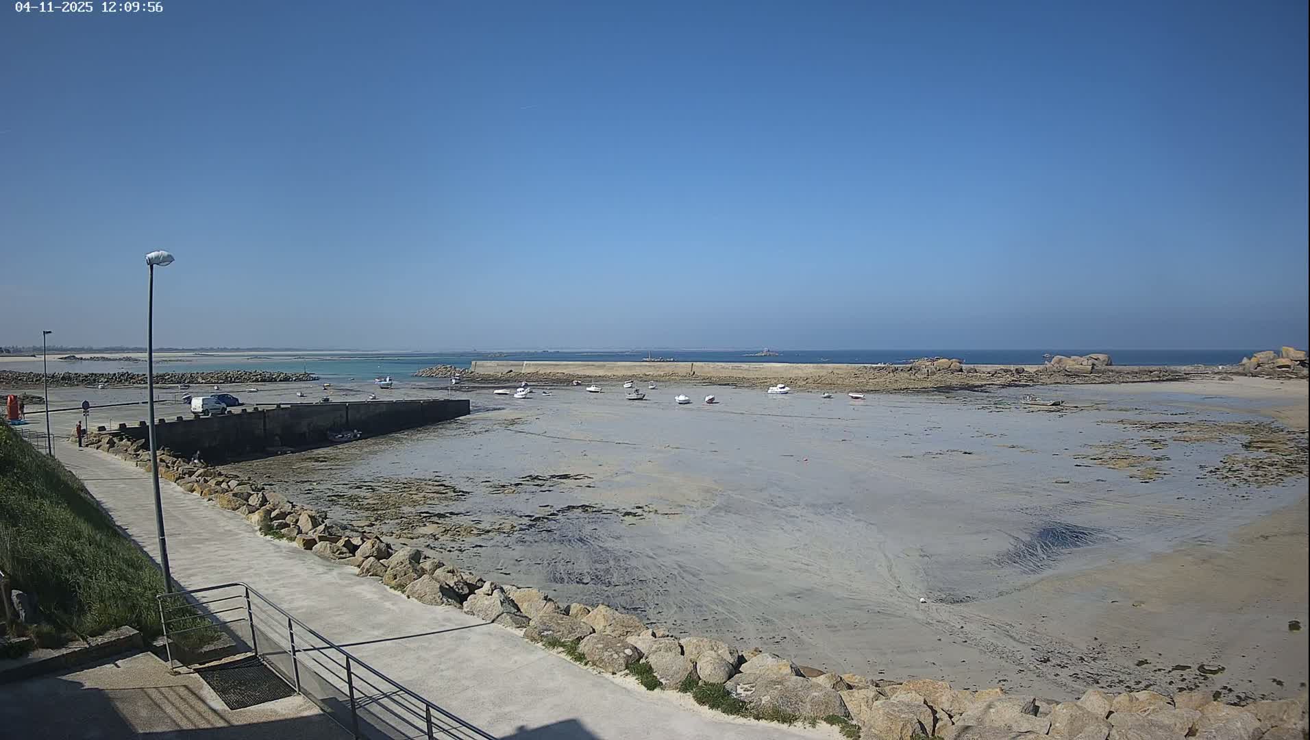 A mostly sunny day reveals a small harbor with several boats resting on exposed mudflats at low tide, surrounded by rocky shores and calm ocean waters extending to a distant horizon.