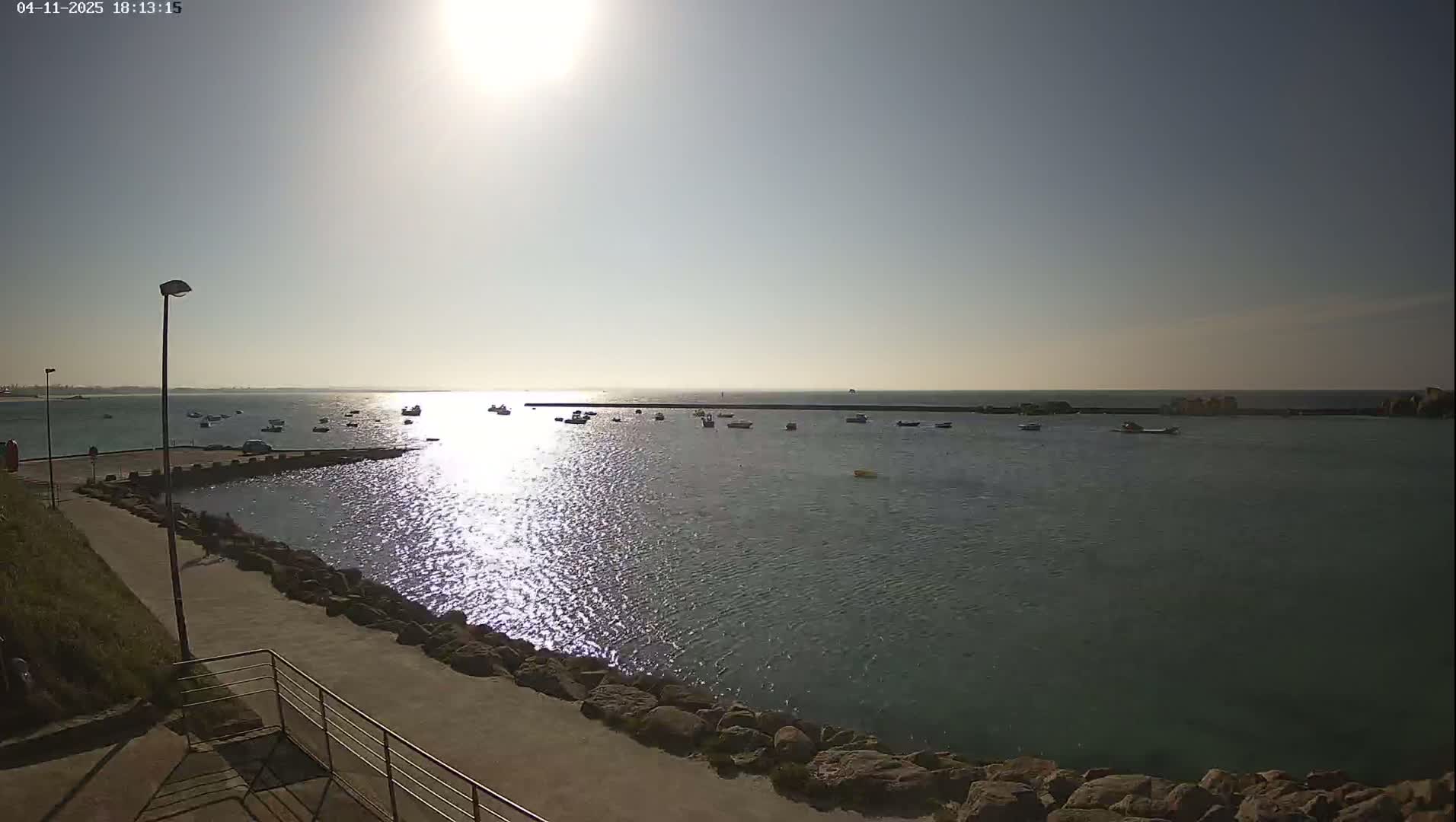 A sunny day at a harbor shows calm water with several small boats moored, a paved walkway leading to a breakwater, and a bright sun in the sky.