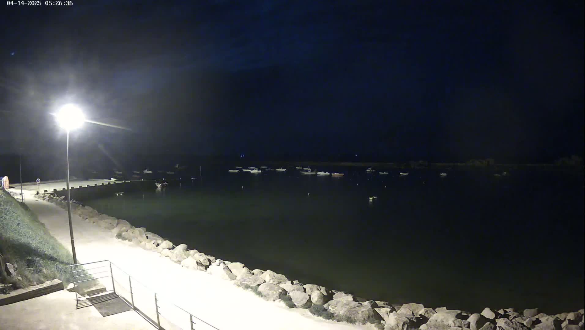 A nighttime view of a calm body of water with several boats moored, illuminated by a single lamppost on a paved walkway beside a rocky shoreline.