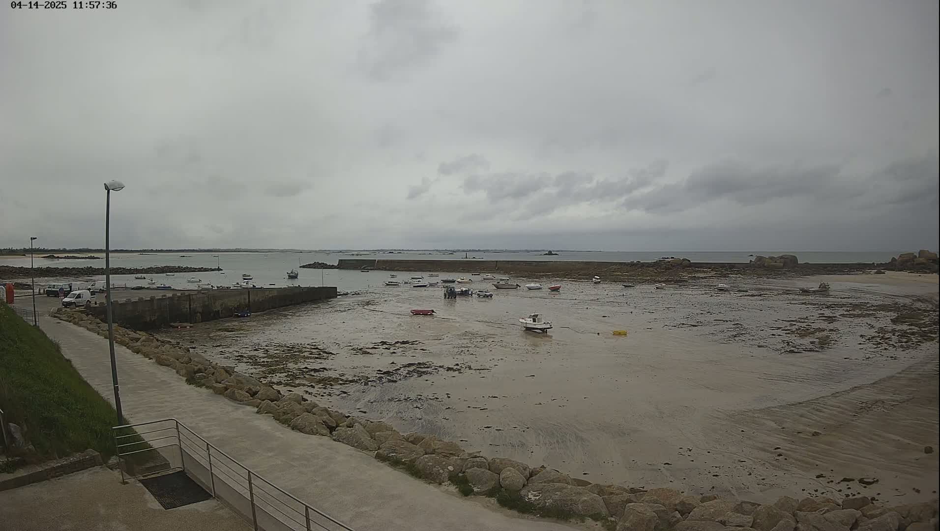 A mostly overcast day reveals a small harbor with numerous small boats resting on exposed mudflats, a calm sea beyond a low stone wall, and a paved pathway leading down to the water.