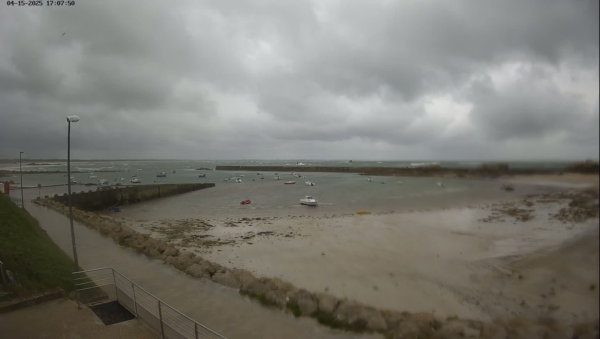 A small harbor with several boats is mostly exposed at low tide under an overcast sky, with choppy waves visible in the distance.