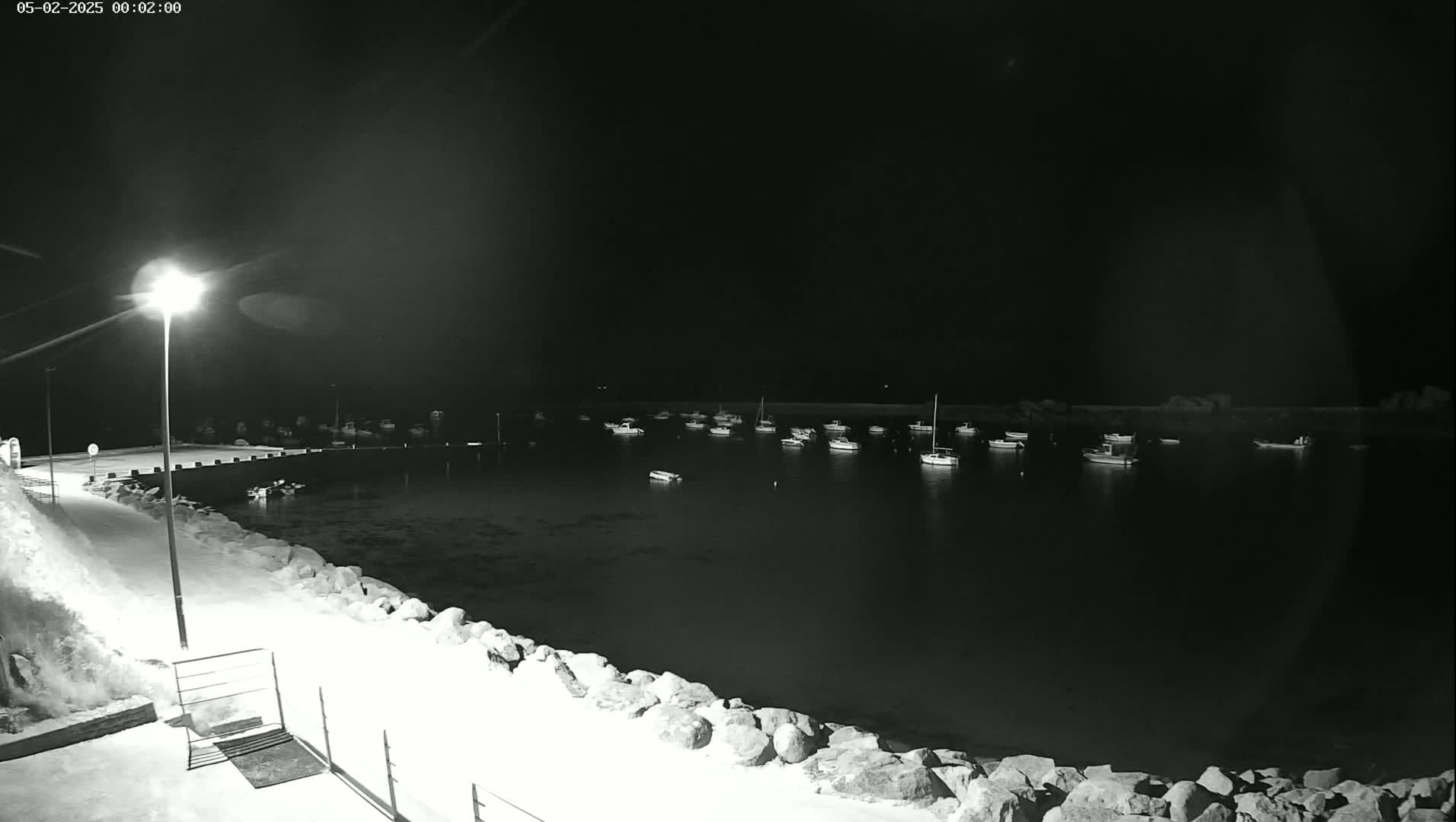 A nighttime view of a harbor with numerous boats at anchor, illuminated by a single lamppost on a snow-covered dock.