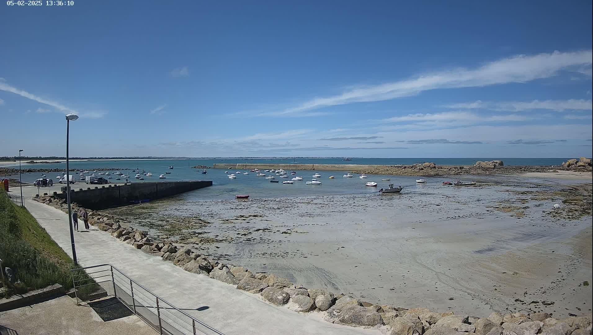 A mostly sunny day reveals a small harbor with numerous boats at low tide, a paved walkway leading to it, and a calm sea beyond.