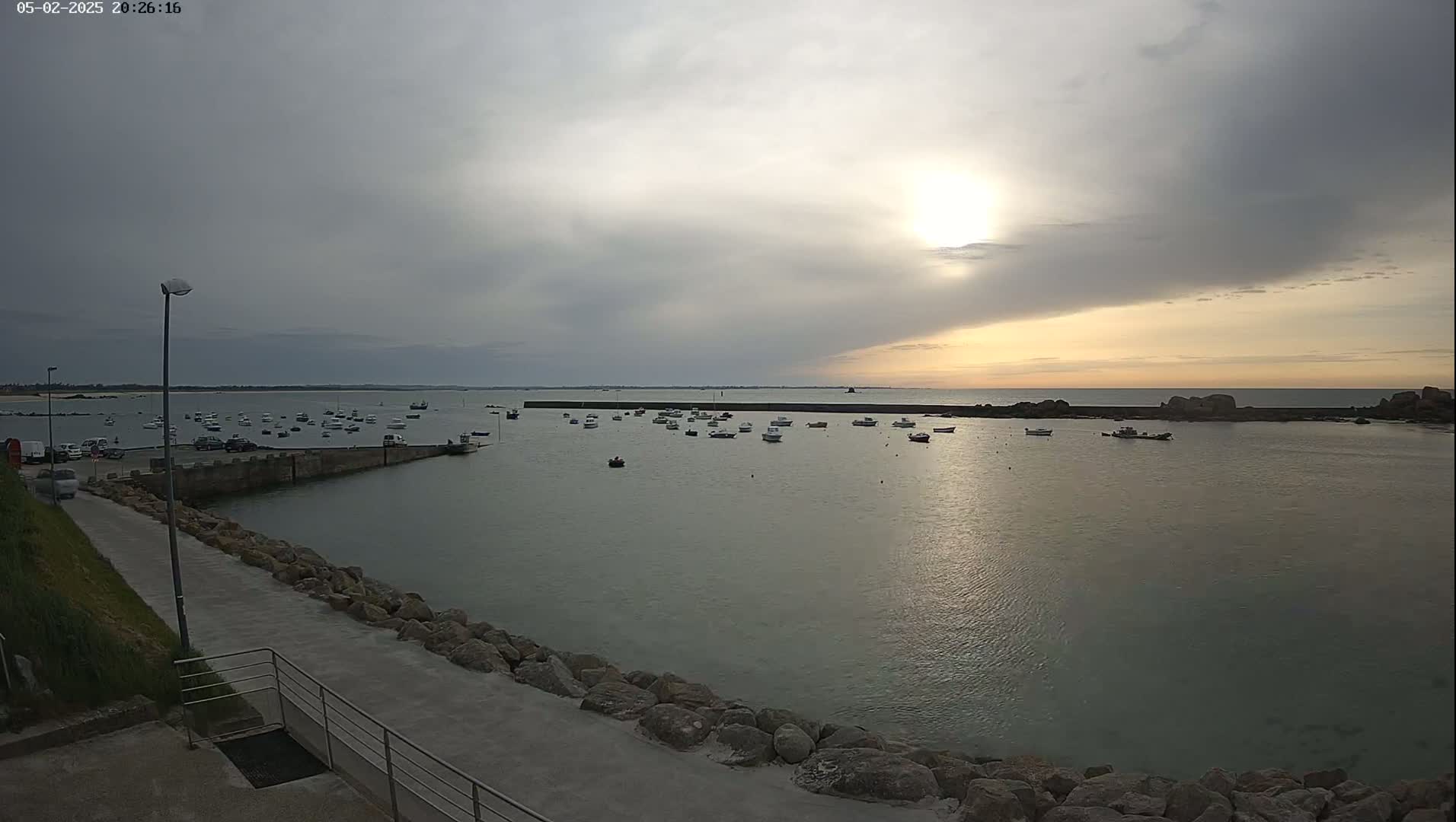 A calm harbor filled with numerous small boats is seen under an overcast sky with a bright sun partially visible.