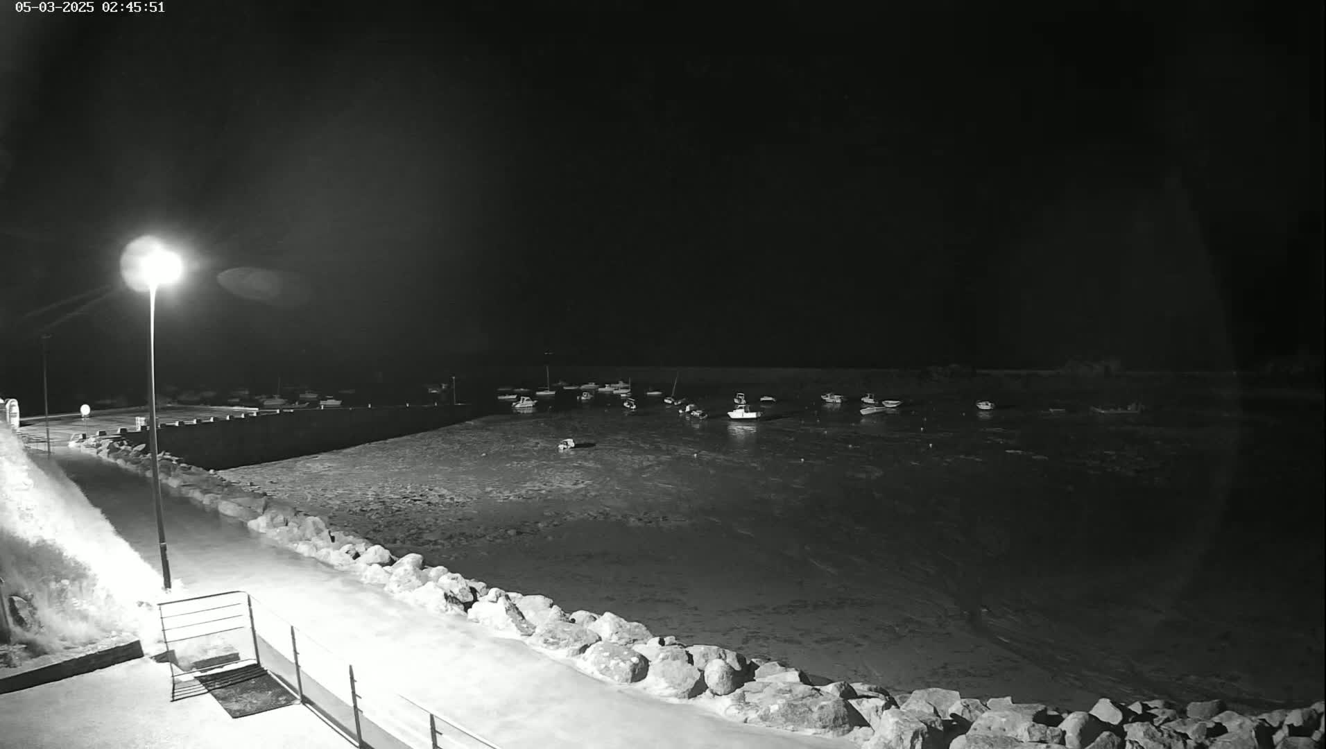 A nighttime view of a harbor filled with boats, partially frozen water, and a snow-covered walkway lit by a single lamppost.