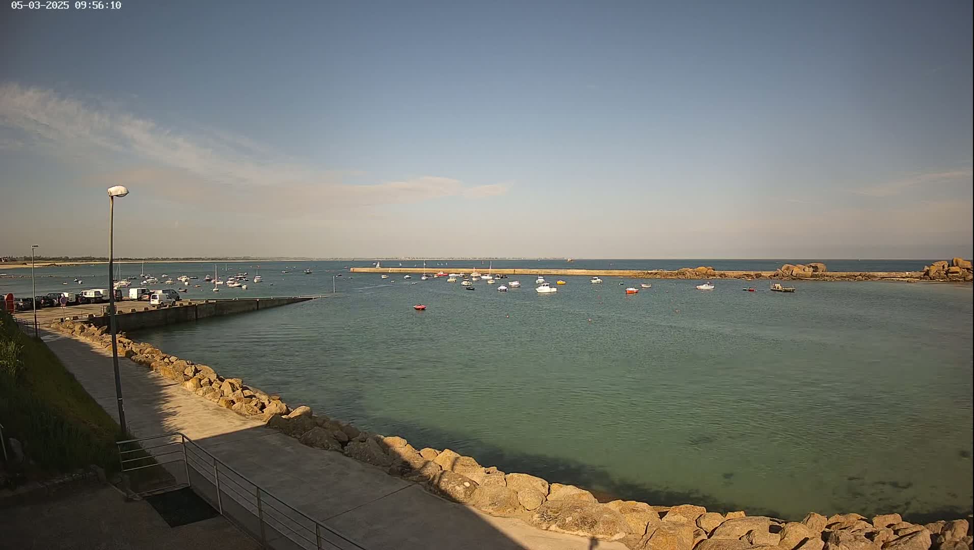 A calm, sunny day reveals a harbor filled with small boats, a rocky shoreline, and a parking lot with several vehicles.