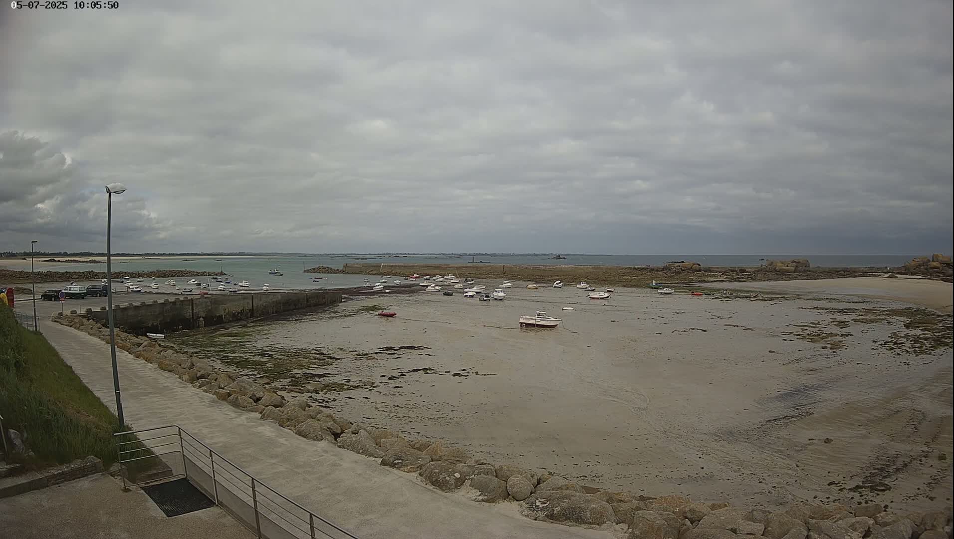 A mostly overcast sky overlooks a harbor filled with small boats on a low tide, with a paved walkway and grassy area in the foreground.