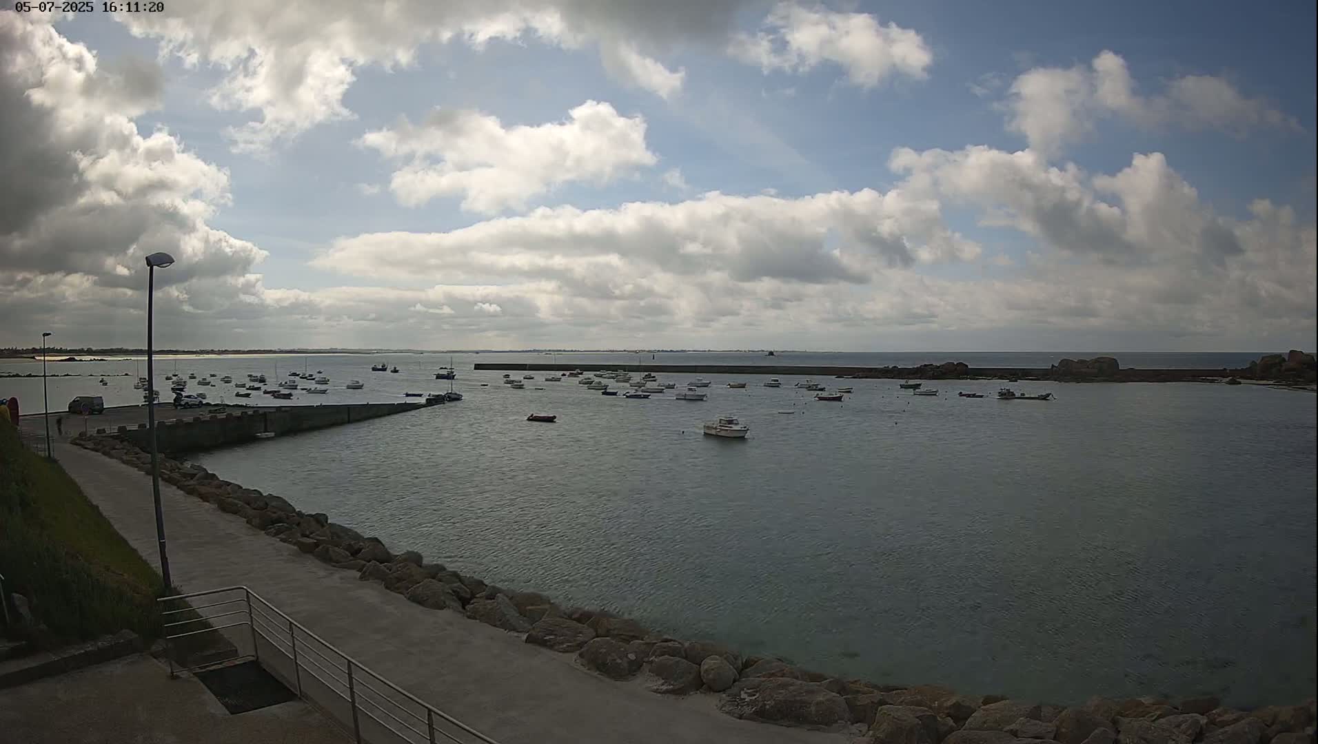 A partly cloudy day shows numerous small boats moored in a calm harbor, with a paved walkway and rock barrier leading to the water.