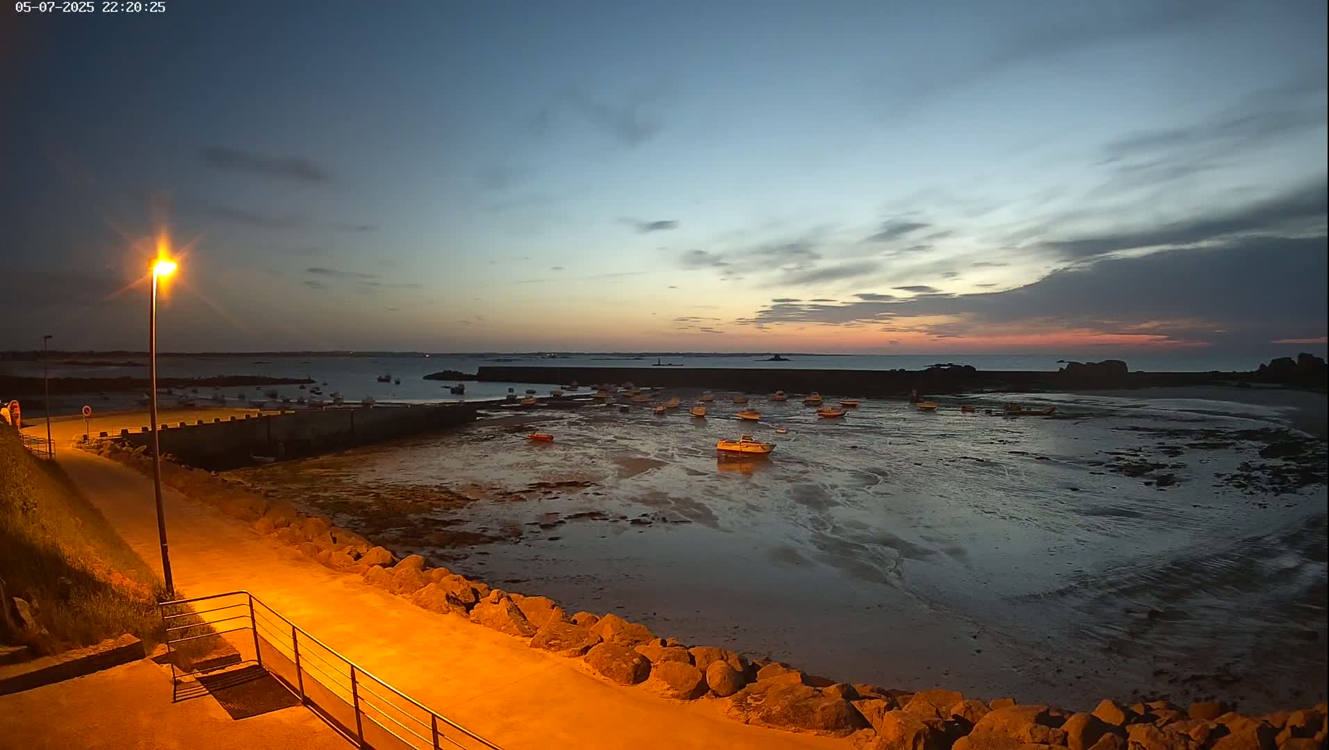 A calm, twilight-lit harbor scene shows numerous small boats resting on exposed mudflats at low tide, under a partly cloudy sky.