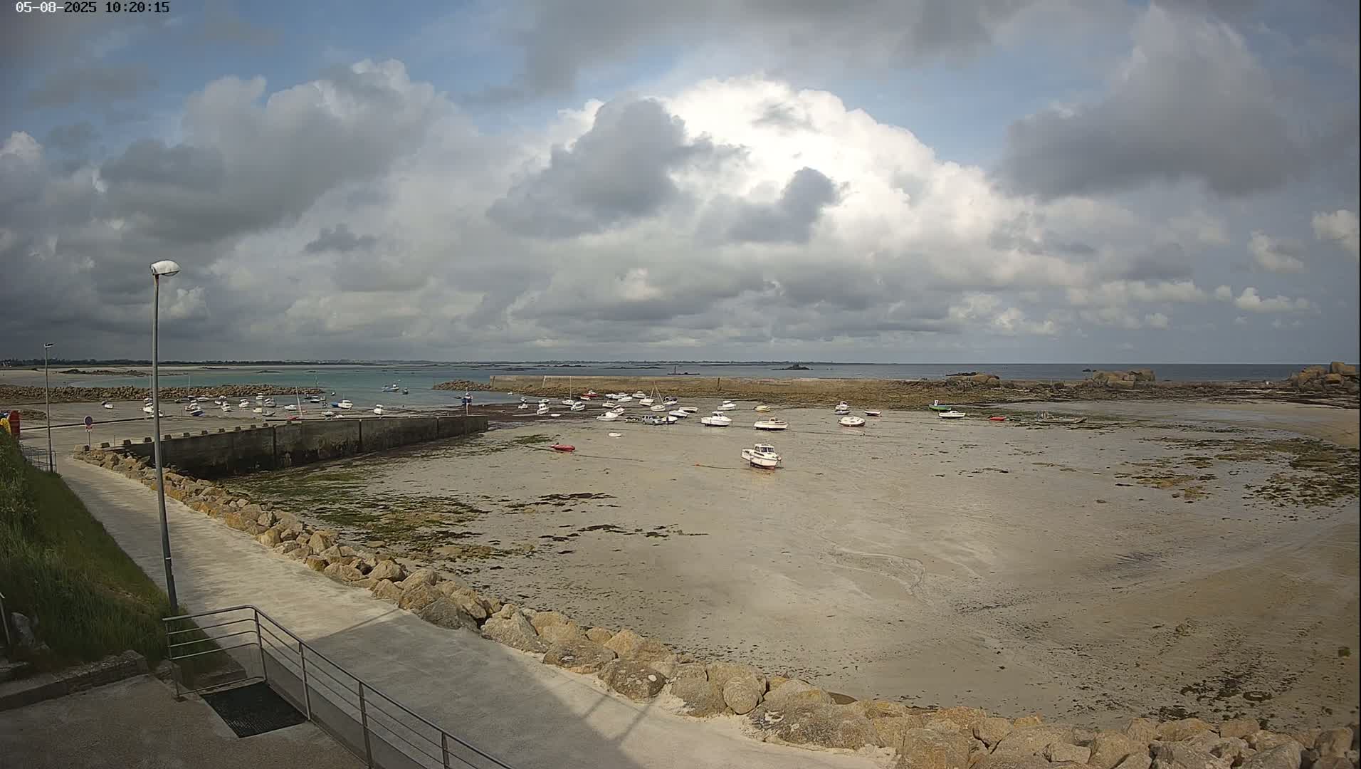 A mostly cloudy day reveals a sandy, low-tide harbor with numerous small boats resting on the exposed seabed, a stone seawall, and a paved walkway leading towards the viewer.