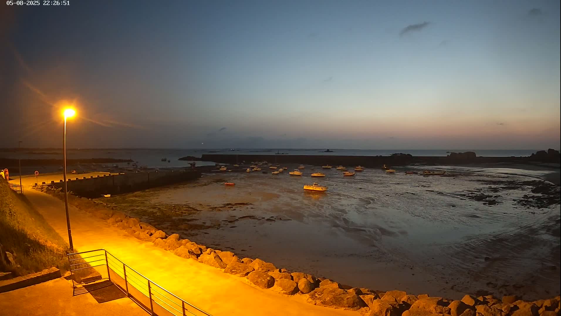 A low-lit harbor at twilight, showing several small boats resting on the exposed seabed at low tide, with a calm sea and a clear sky.