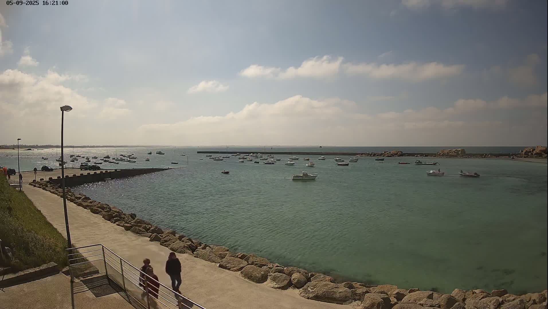 A calm, sunny day shows a harbor filled with numerous small boats, with two people walking along a paved path beside a rocky seawall.