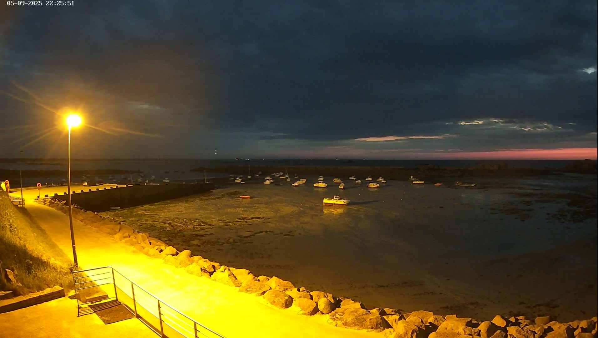 A nighttime view of a harbor filled with small boats under a partly cloudy sky, with a pinkish hue on the horizon.