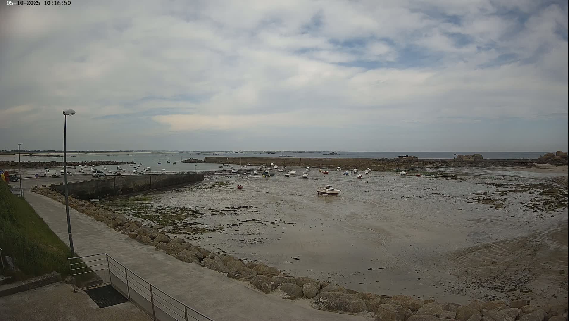 A mostly cloudy day reveals a small harbor with numerous boats resting on exposed mudflats at low tide, a calm sea stretching to a distant horizon.