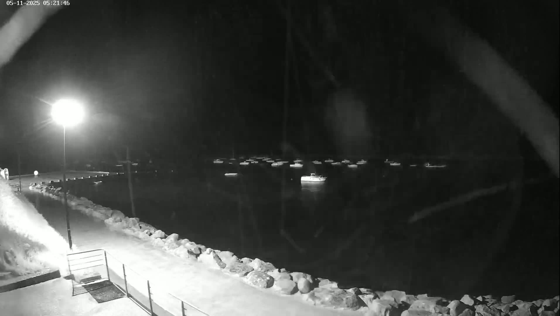 A nighttime, snow-covered waterfront shows numerous boats moored in calm water under a bright streetlight.