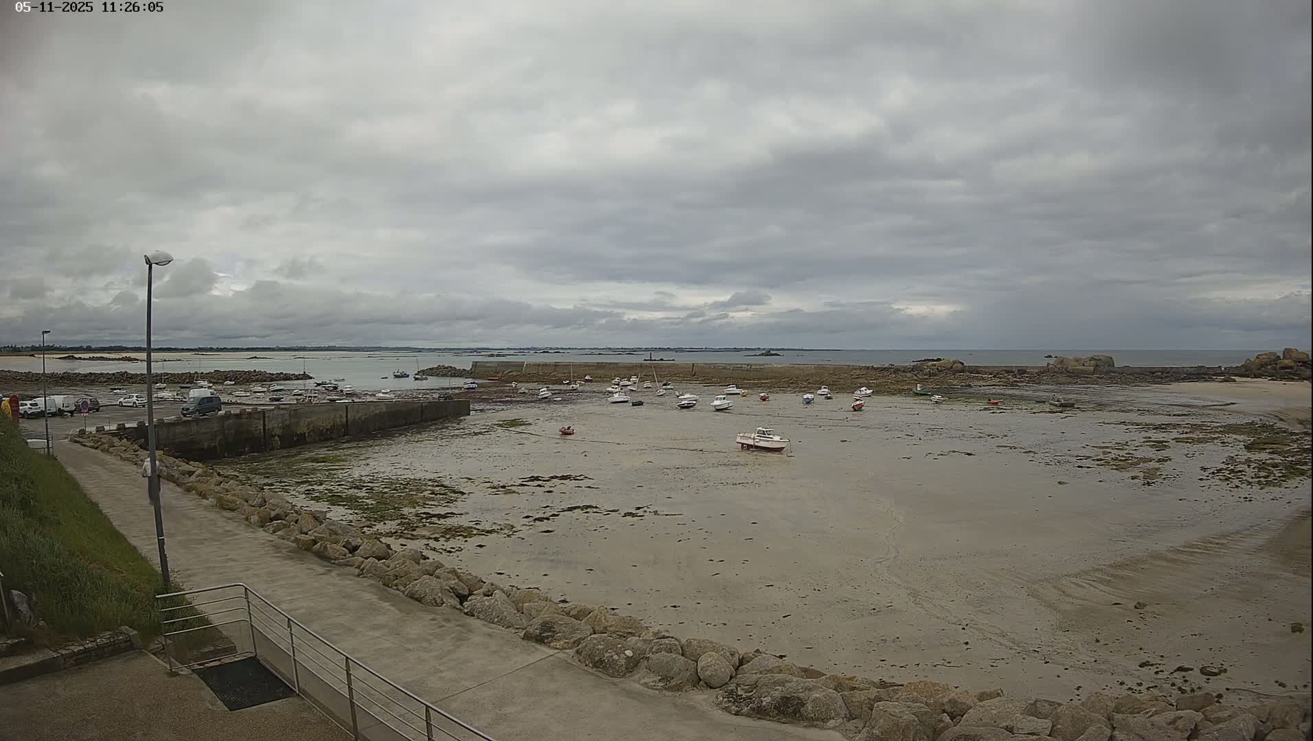 A mostly cloudy day reveals a rocky shoreline with several small boats moored in shallow, sandy water, next to a paved walkway.