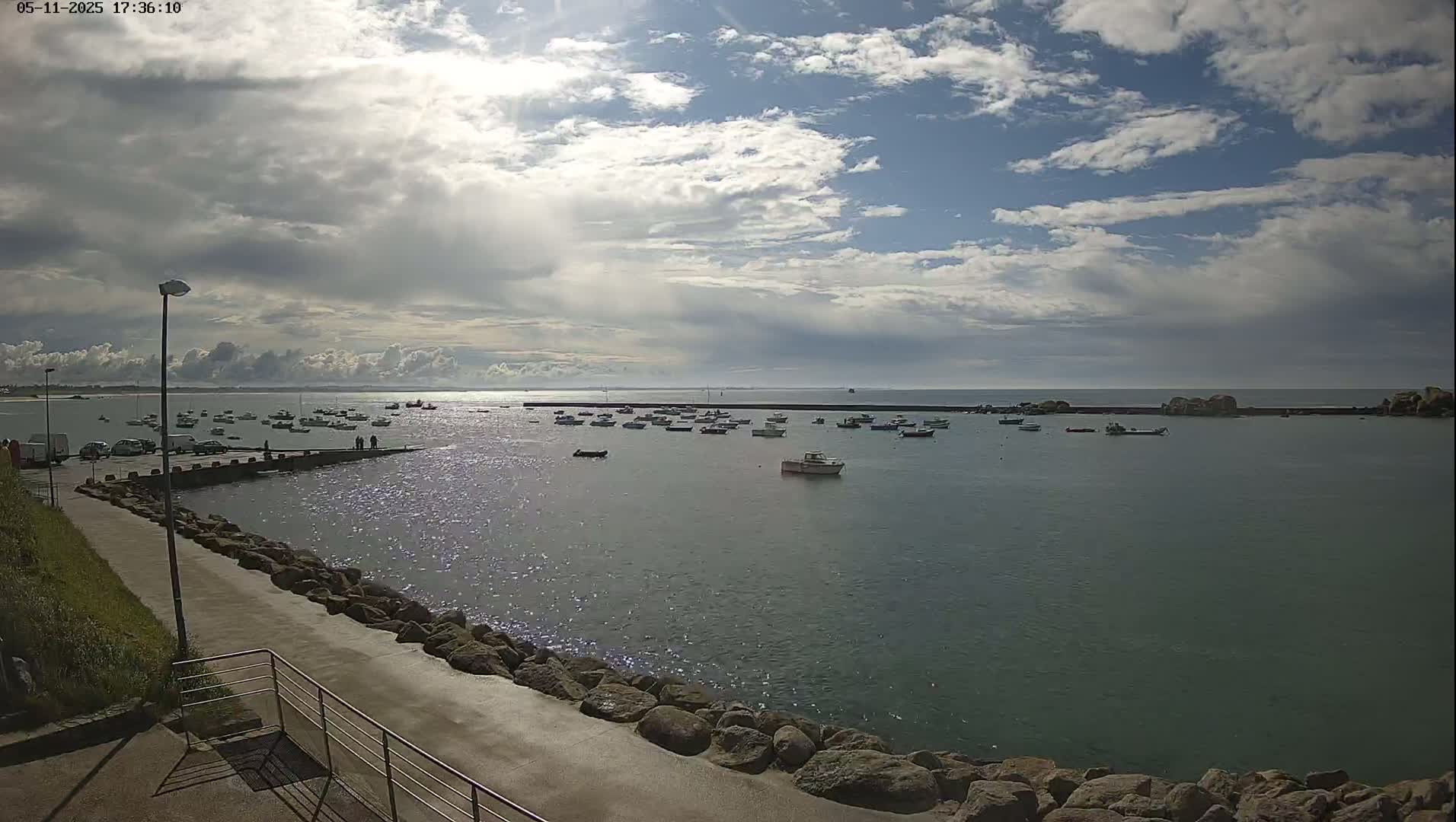 A partly cloudy sky overlooks a calm harbor filled with numerous small boats moored near a paved walkway lined with rocks.