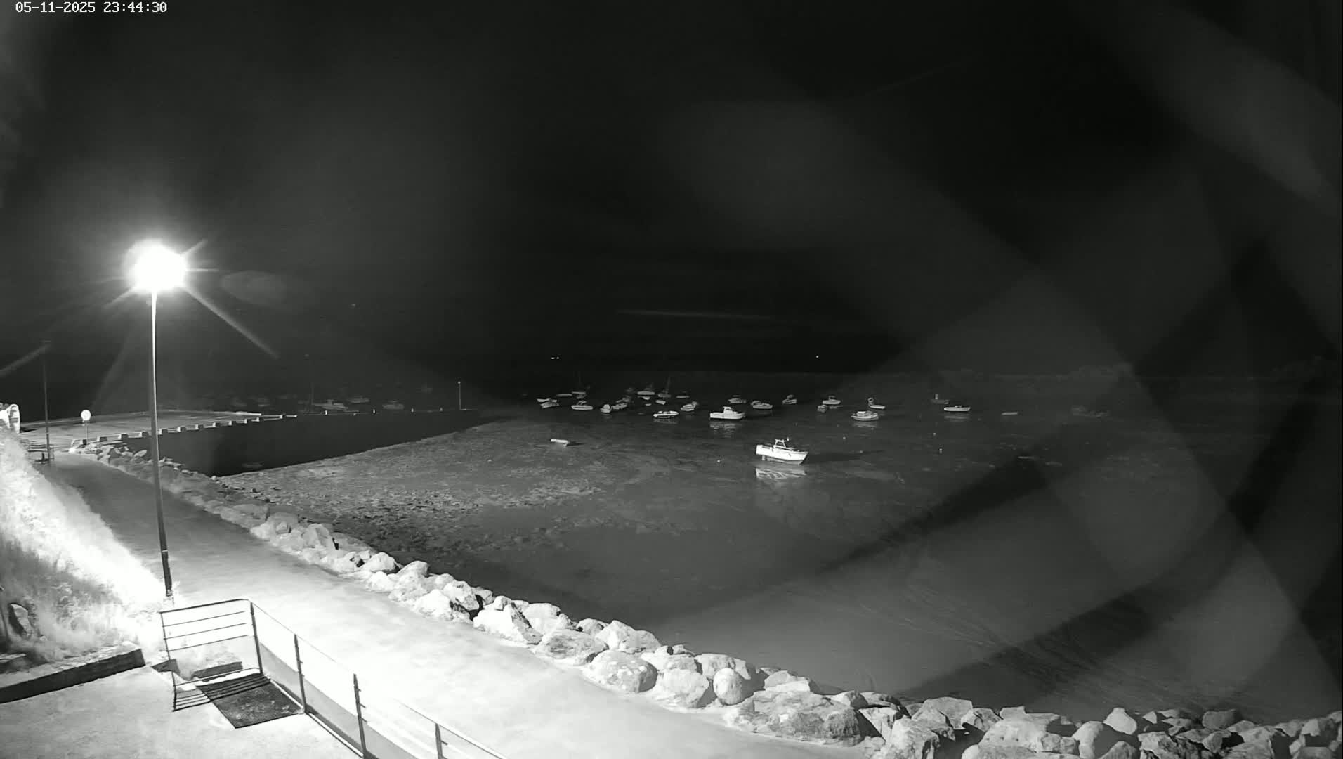 A nighttime, black and white view shows a calm body of water with several boats moored, seen from a paved walkway next to a low stone wall, illuminated by a single streetlight.