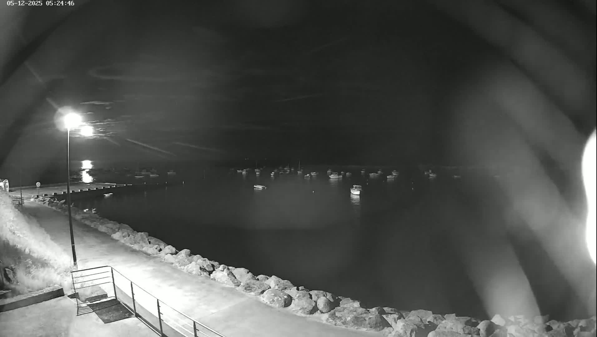 A nighttime view of a calm harbor with numerous boats at anchor, illuminated by a single lamppost under a partly cloudy sky.