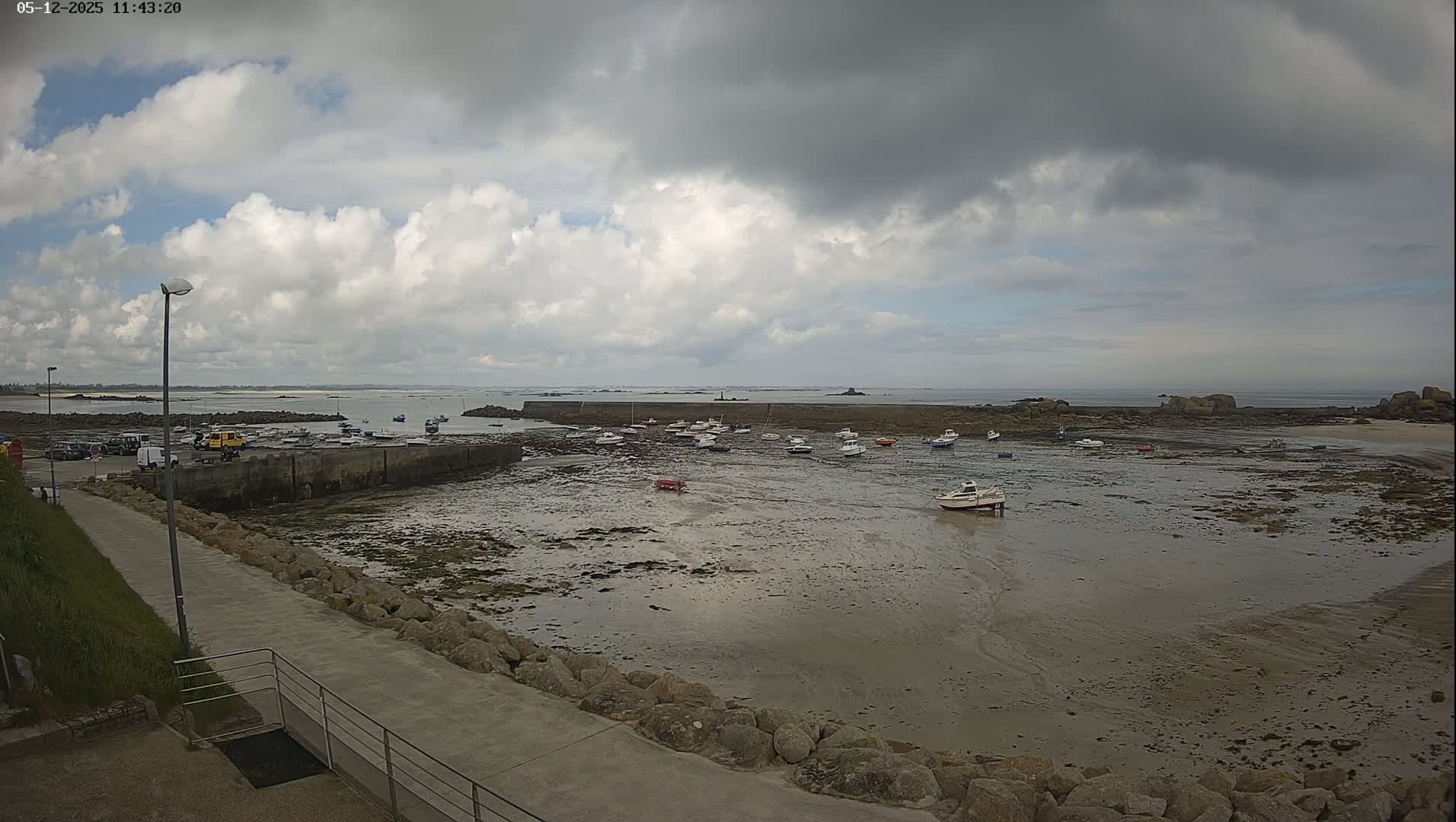 A partly cloudy day reveals a rocky shoreline with numerous small boats resting in shallow, muddy water, next to a paved walkway.