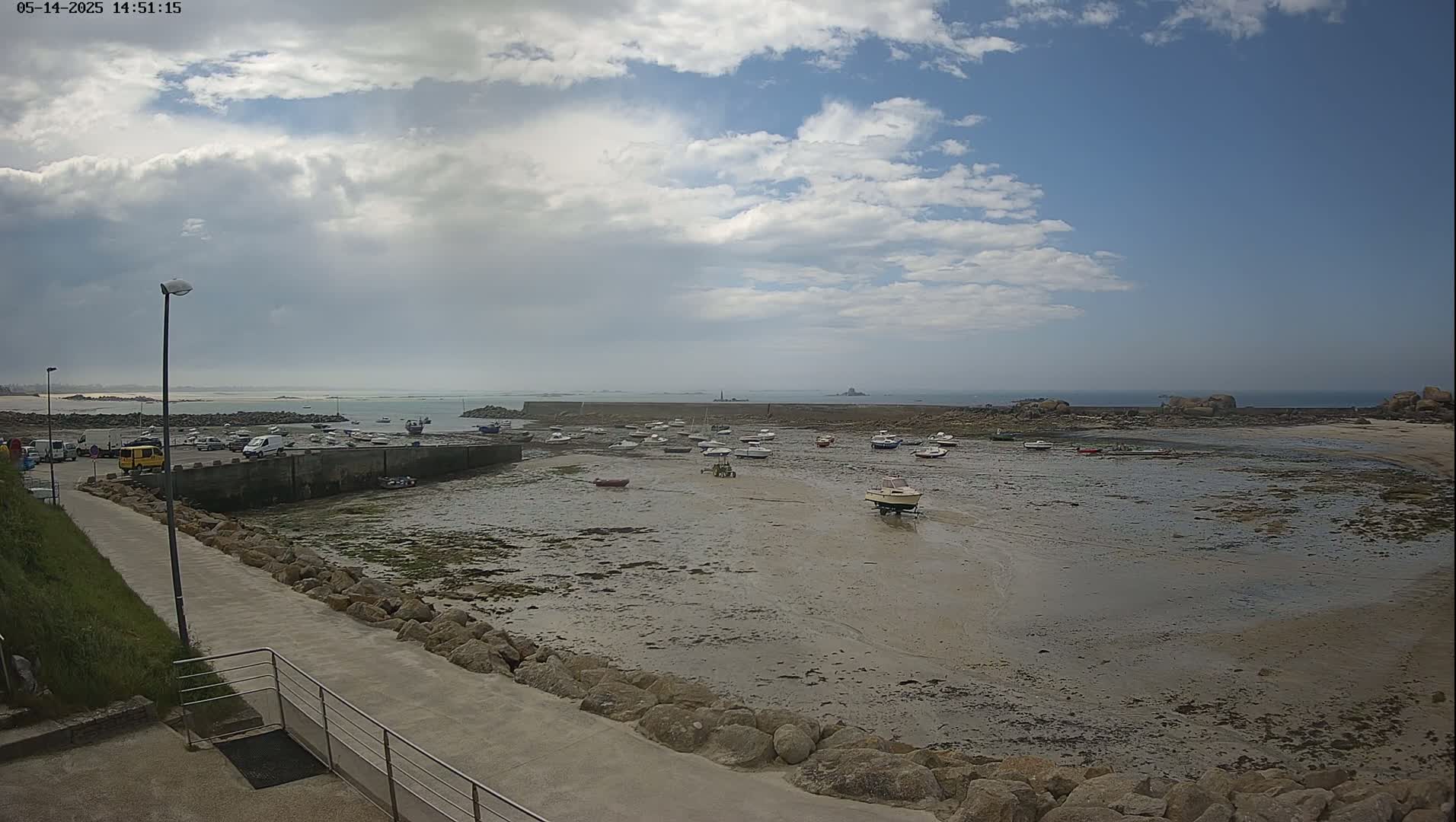 A mostly sunny day shows a harbor with numerous small boats resting on exposed mudflats, a paved walkway leading away, and a calm ocean in the background.