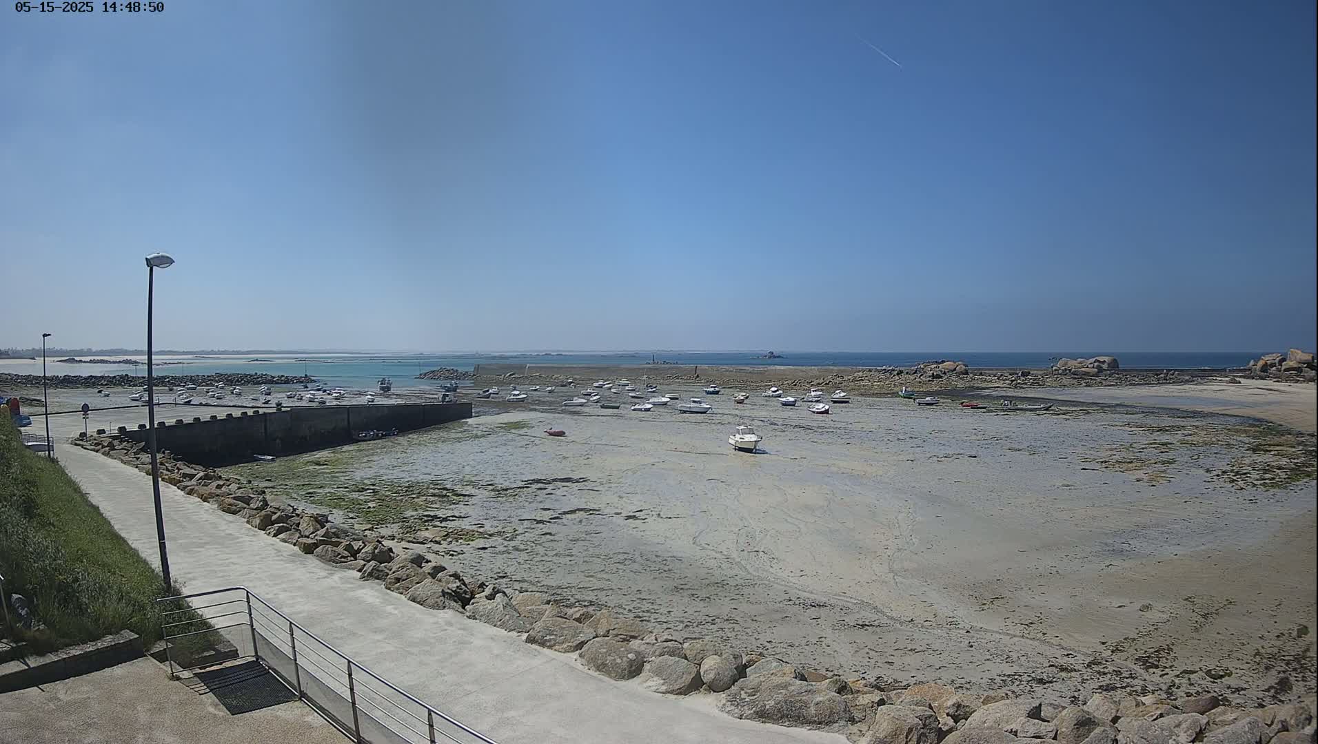A sunny day reveals a mostly exposed harbor filled with numerous small boats at low tide, a paved walkway leads to the water's edge.