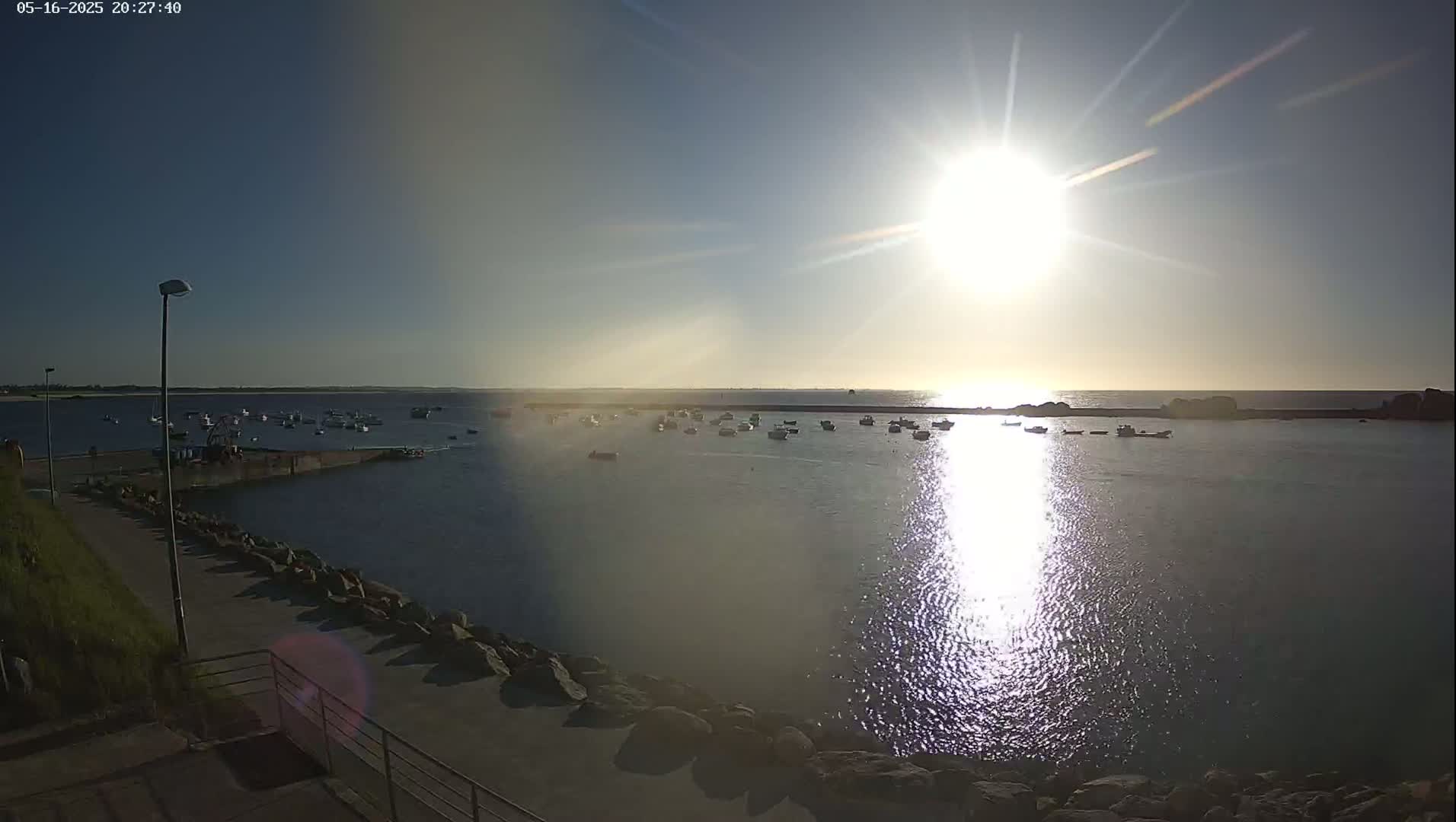 A bright sun shines over a calm harbor filled with numerous small boats, a paved walkway and rocky shoreline visible in the foreground under a clear blue sky.