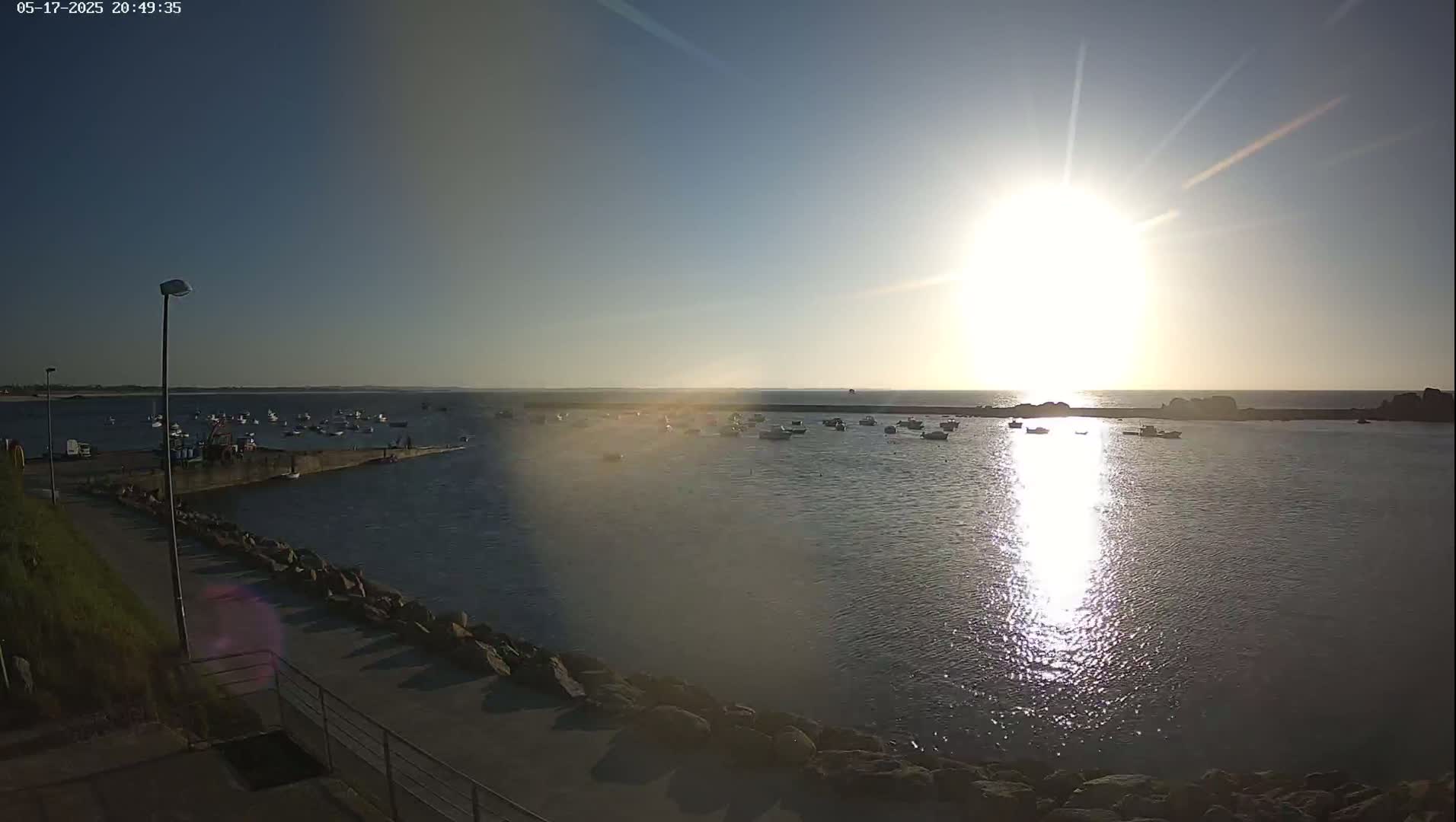 A calm body of water with numerous small boats is seen at sunset, under a bright, sunny sky.