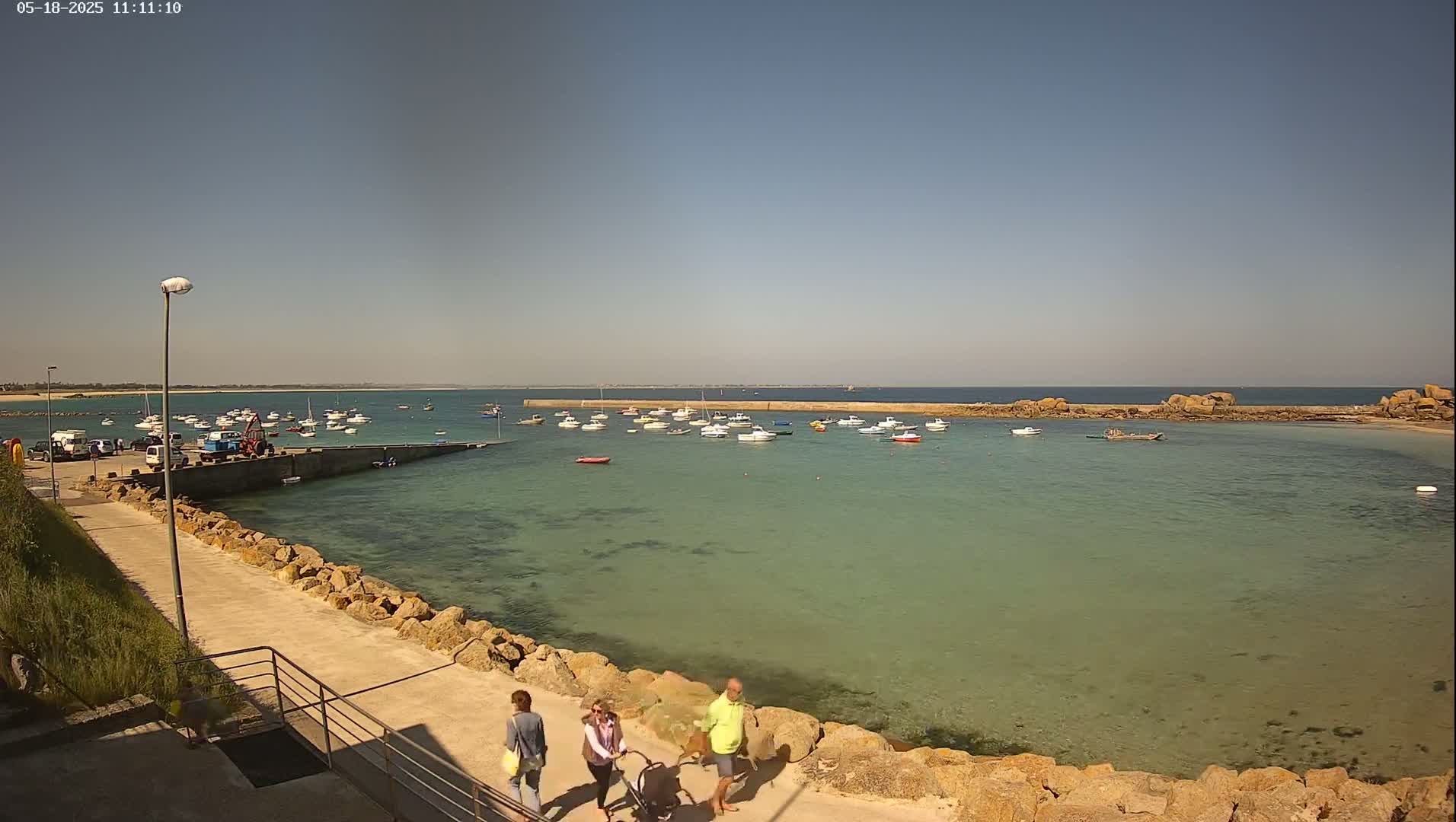 A calm, sunny day reveals a small harbor filled with numerous boats, with a few people strolling along a stone walkway overlooking the clear, shallow water.