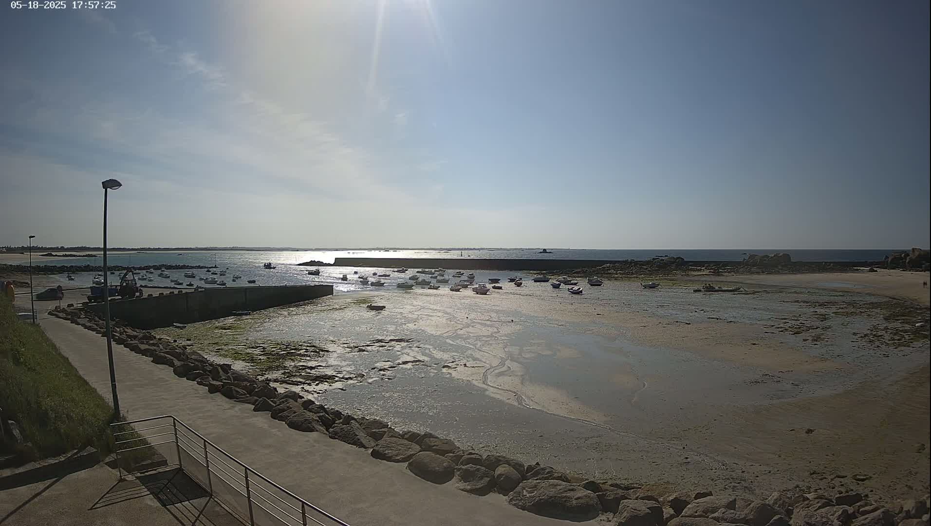 A sunny day at a harbor shows numerous small boats resting on a sandy, low-tide beach, with a walkway leading to the water.