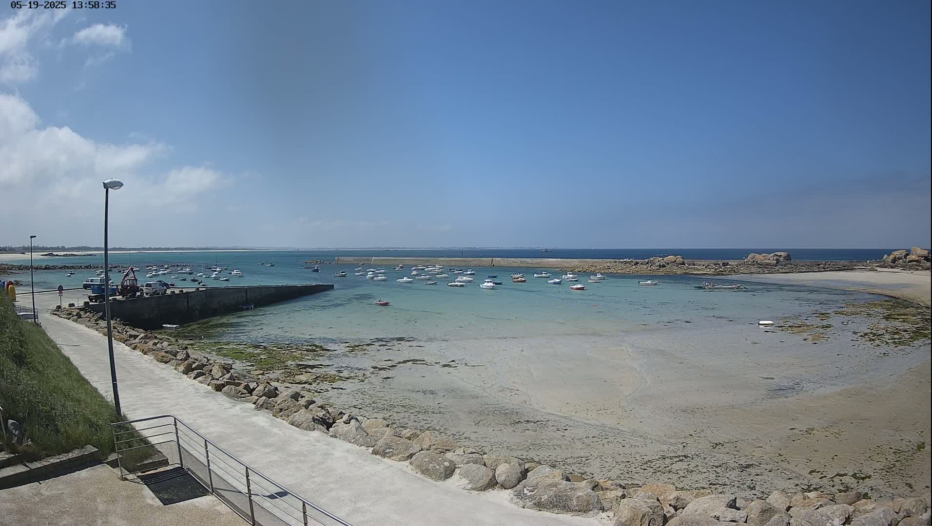 A sunny day reveals a calm harbor filled with numerous small boats, a rocky shore, and a paved walkway leading to the water.